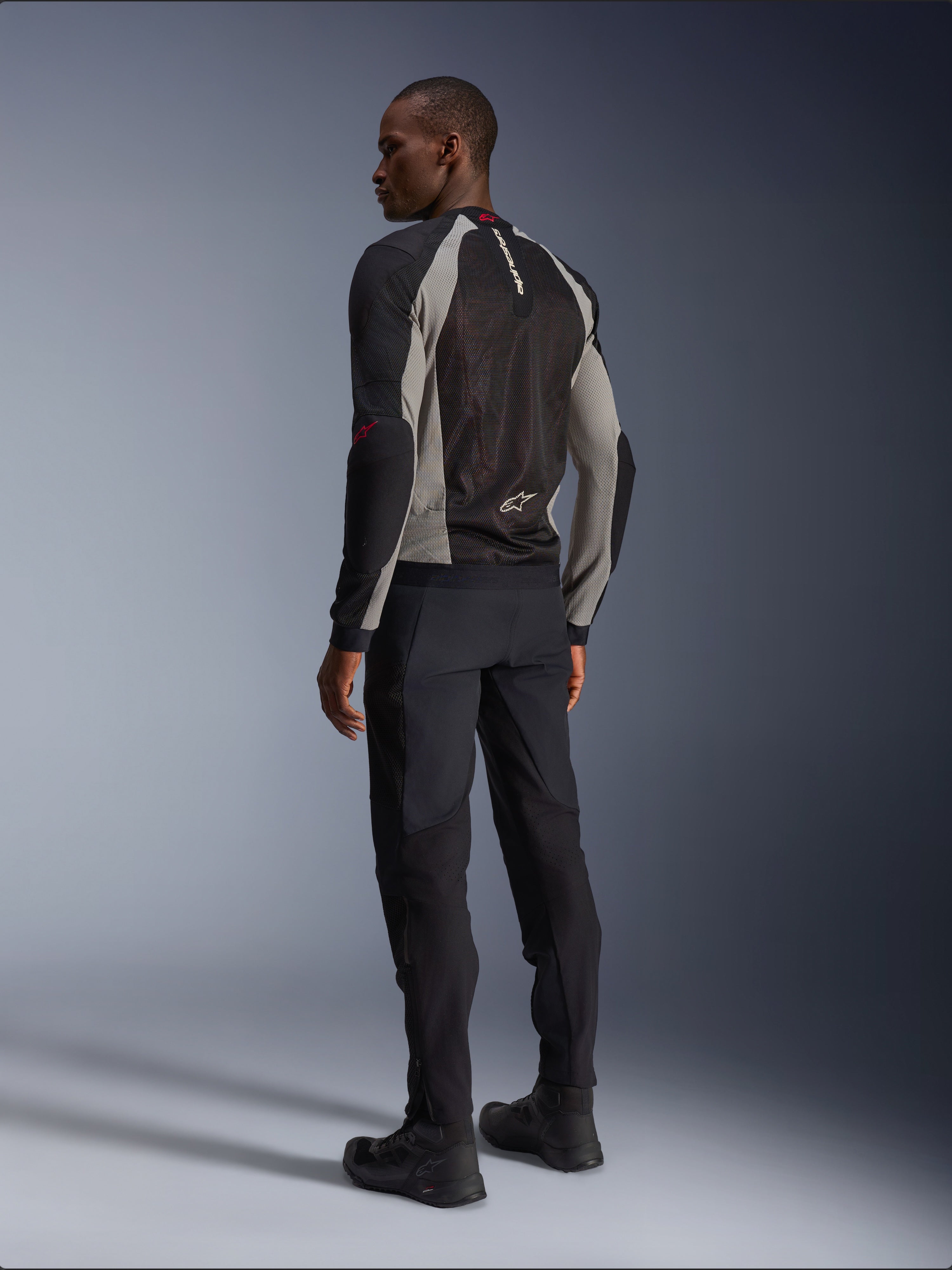 Model wearing Alpinestars Troop-Air Jacket from the back, Black Ash Gray and Bright Red accents, technical mesh construction for ventilation, paired with black motorcycle pants and riding boots, urban contemporary style