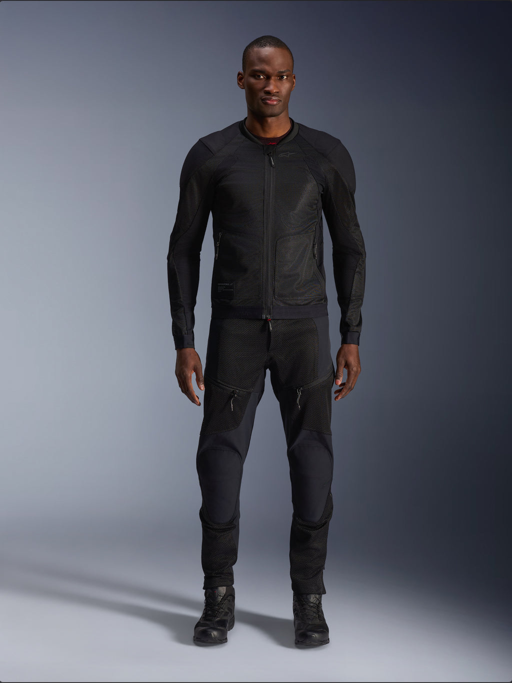 Modelo masculino a usar o Casaco Alpinestars Troop-Air em preto, casaco de motociclismo em malha leve, combinado com calças e botas de condução pretas a condizer, com uma construção técnica em malha 2D para um fluxo de ar superior.
