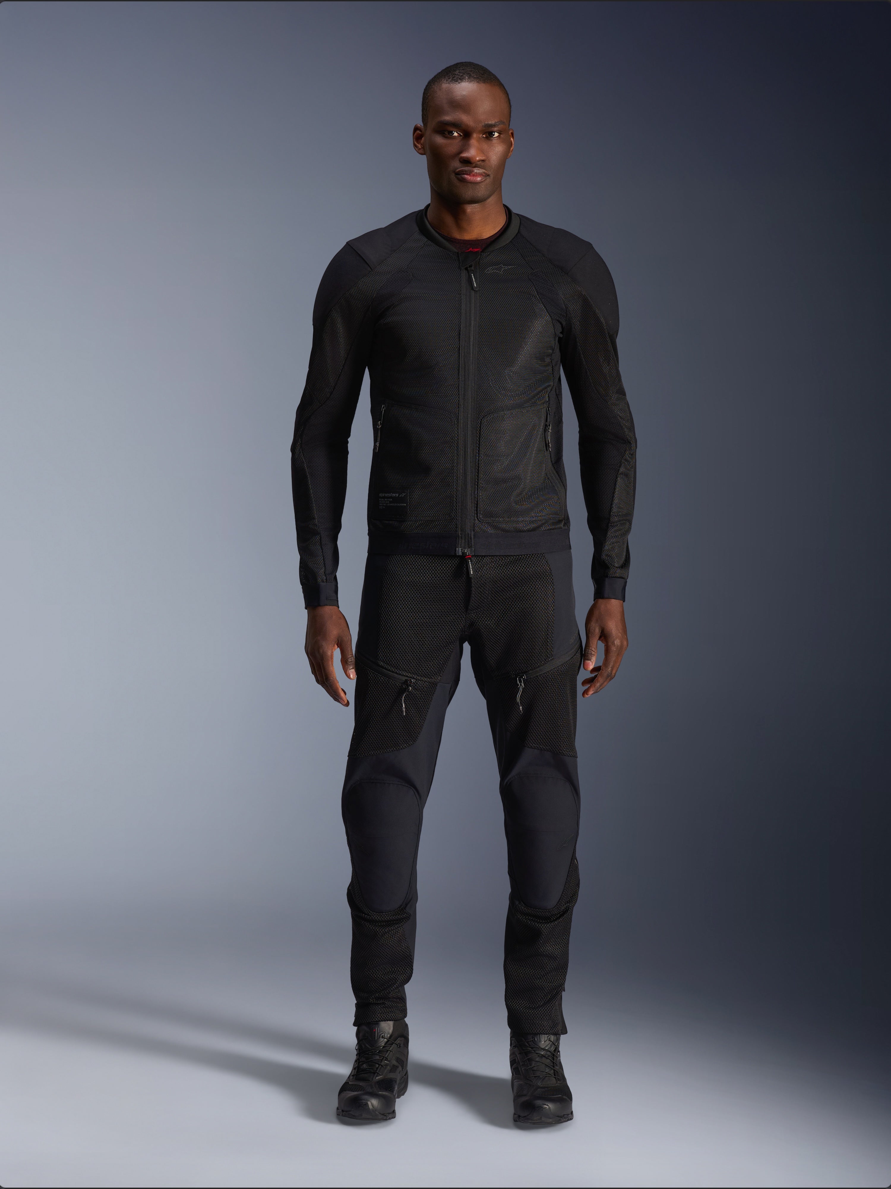Modelo masculino a usar o Casaco Alpinestars Troop-Air em preto, casaco de motociclismo em malha leve, combinado com calças e botas de condução pretas a condizer, com uma construção técnica em malha 2D para um fluxo de ar superior.