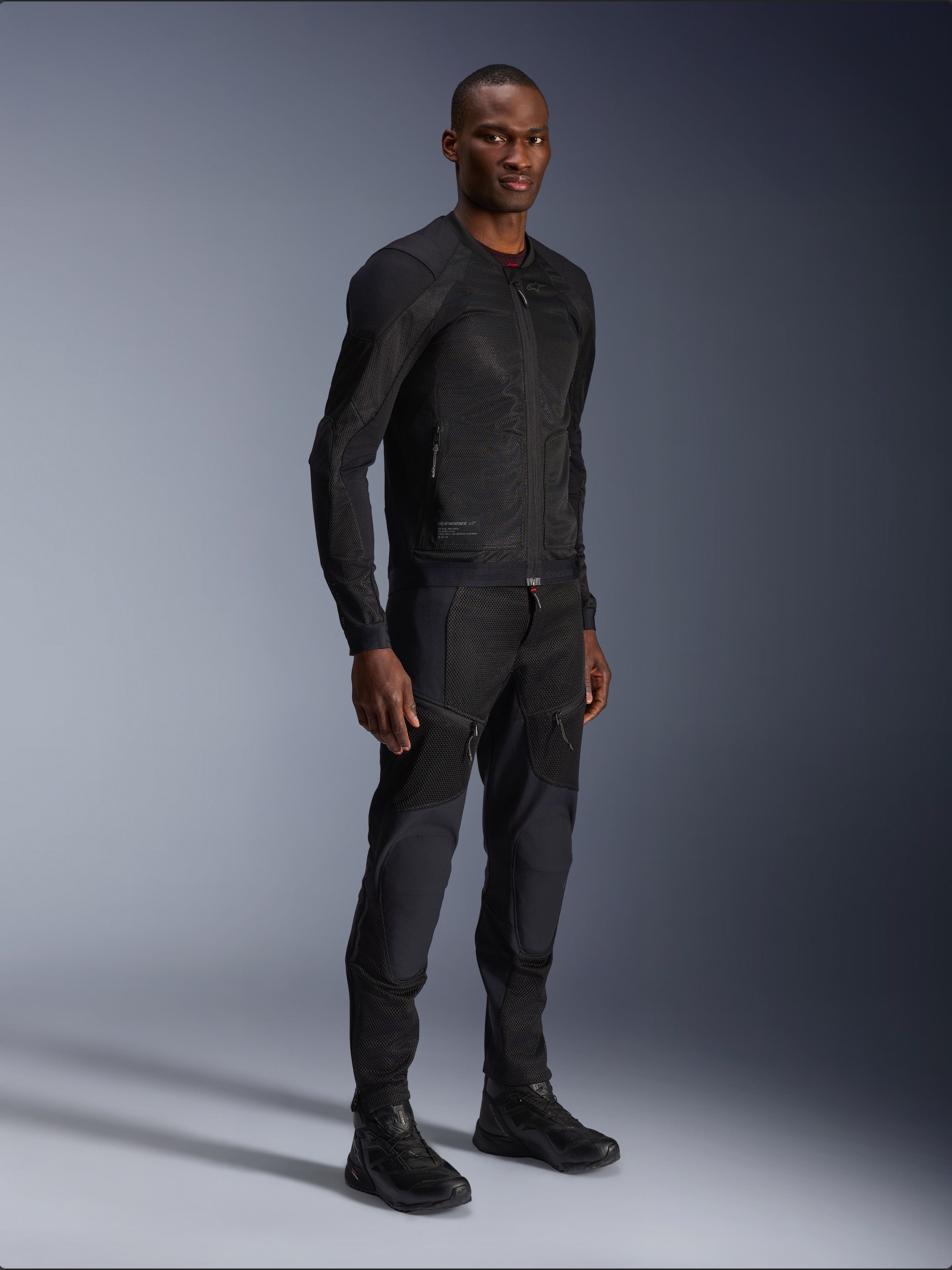 Alpinestars Troop-Air Jacket, casaco de motociclismo em malha técnica, preto, usado por um modelo com calças e botas pretas a condizer, com construção em malha leve e um design urbano minimalista