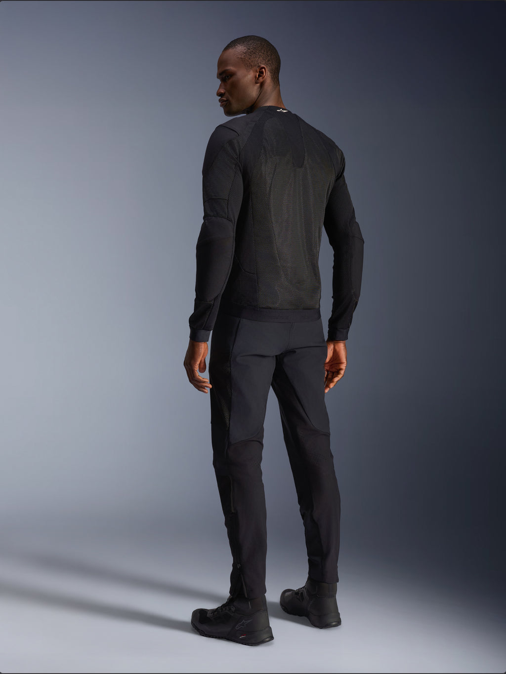 Alpinestars Troop-Air Jacket, casaco de motociclismo técnico preto, vista traseira com amplos painéis de malha para uma ventilação superior, usado com calças pretas e botas de condução, design urbano contemporâneo elegante para condução no verão.