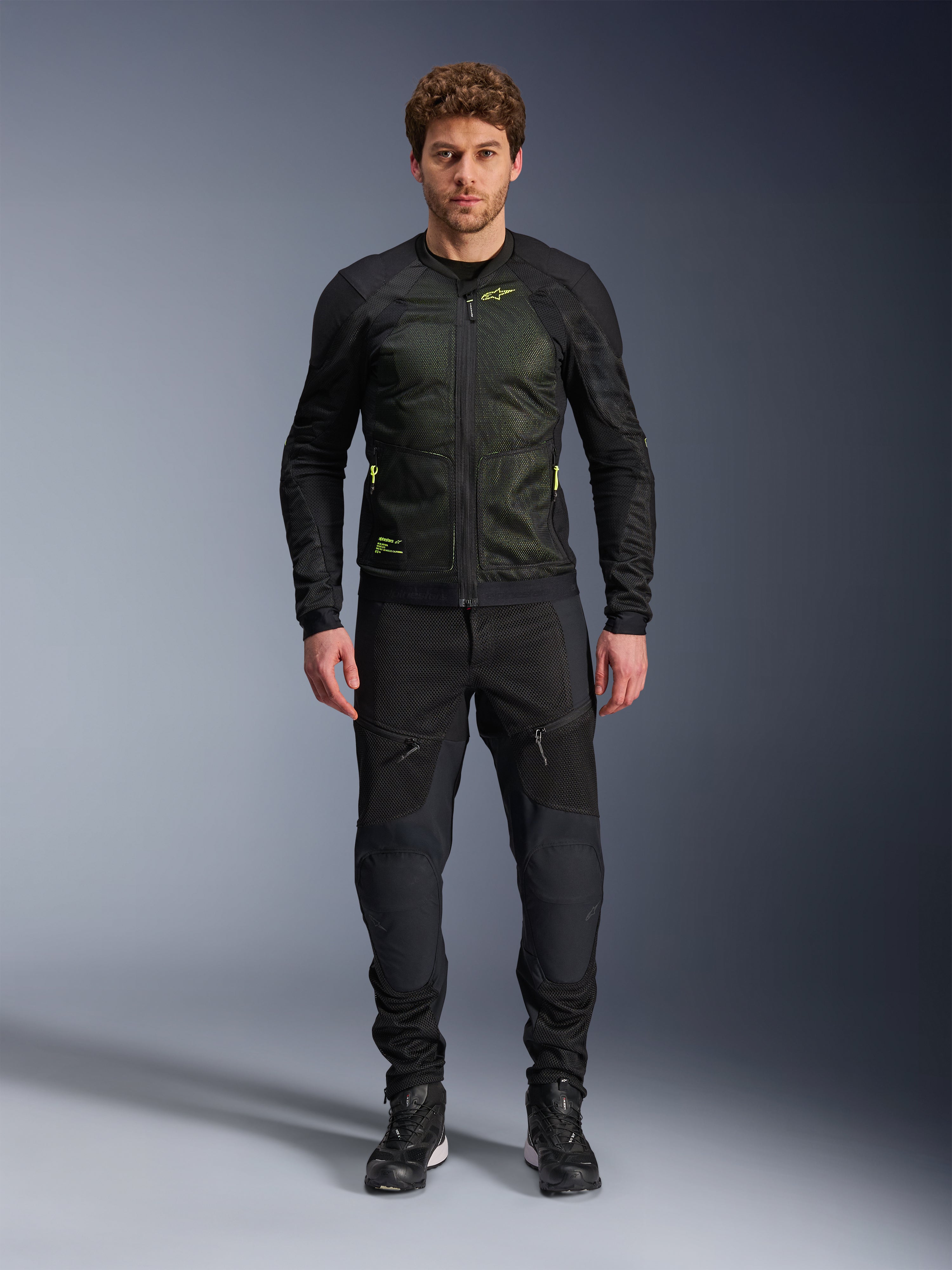 Modelo masculino a usar o Casaco Alpinestars Troop-Air em Preto Amarelo Fluo, com painéis de malha técnica e detalhes em amarelo vibrante, combinado com calças de motociclismo e calçado pretos, vestuário de motociclismo contemporâneo para tempo quente