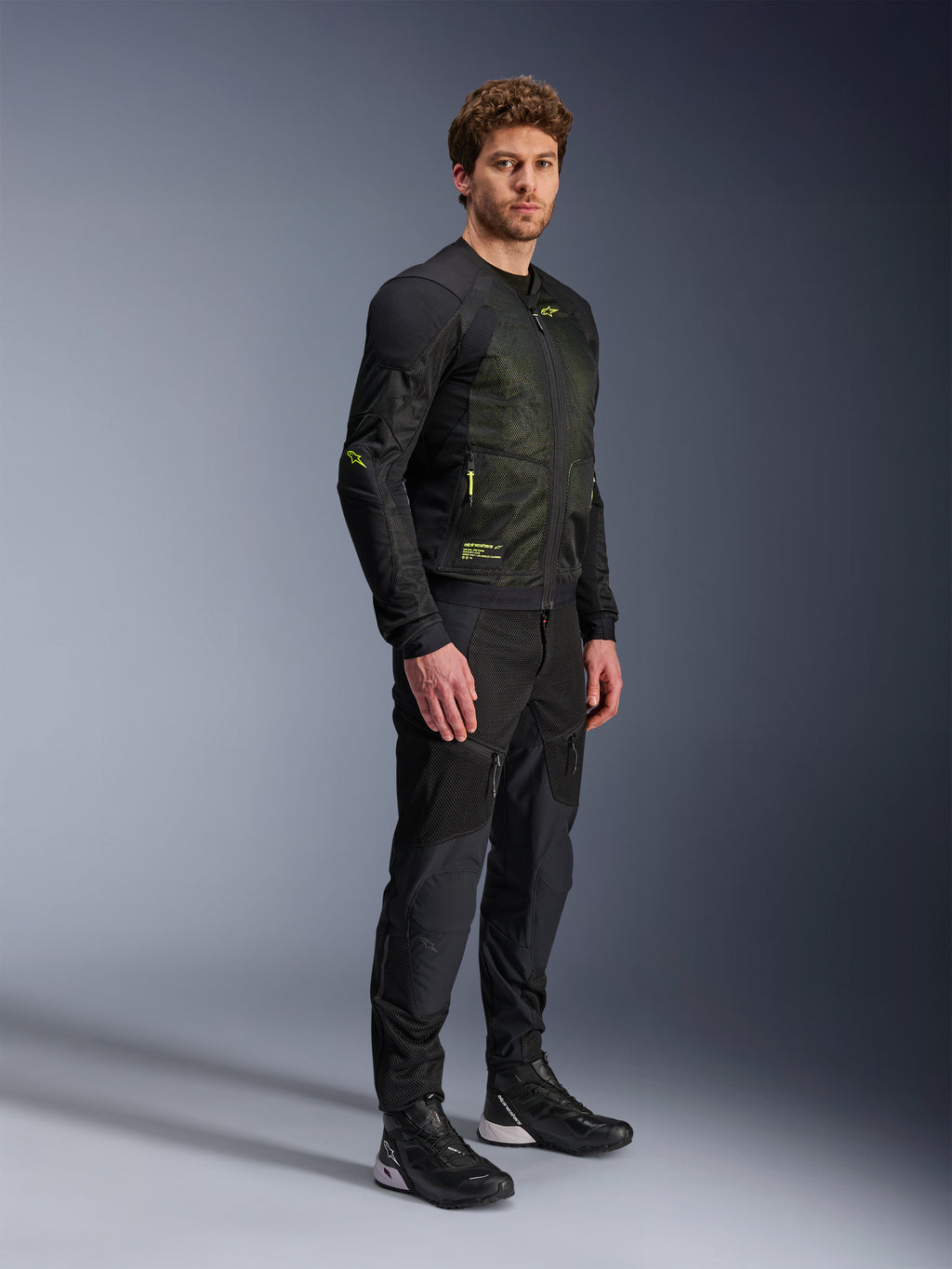 Casaco Alpinestars Troop-Air, casaco de malha para motociclismo, preto e amarelo fluo, vestido por um modelo com calças pretas e sapatilhas de condução a condizer, apresenta construção em malha de elevado fluxo de ar com detalhes em amarelo fluorescente no peito e nas mangas, design técnico leve para condução em estrada
