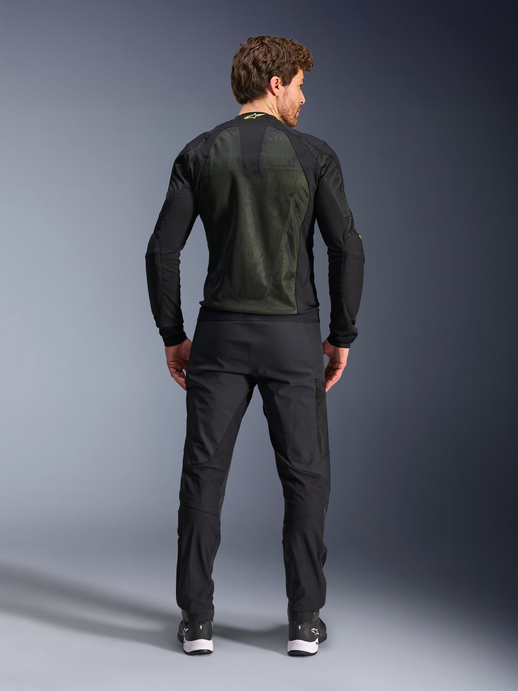 Alpinestars Troop-Air Jacket, Preto Amarelo Fluo, vista traseira num modelo, com um grande painel traseiro em malha técnica 2D para ventilação, casaco técnico de motociclismo usado com calças e botas pretas, estilo urbano de motociclismo