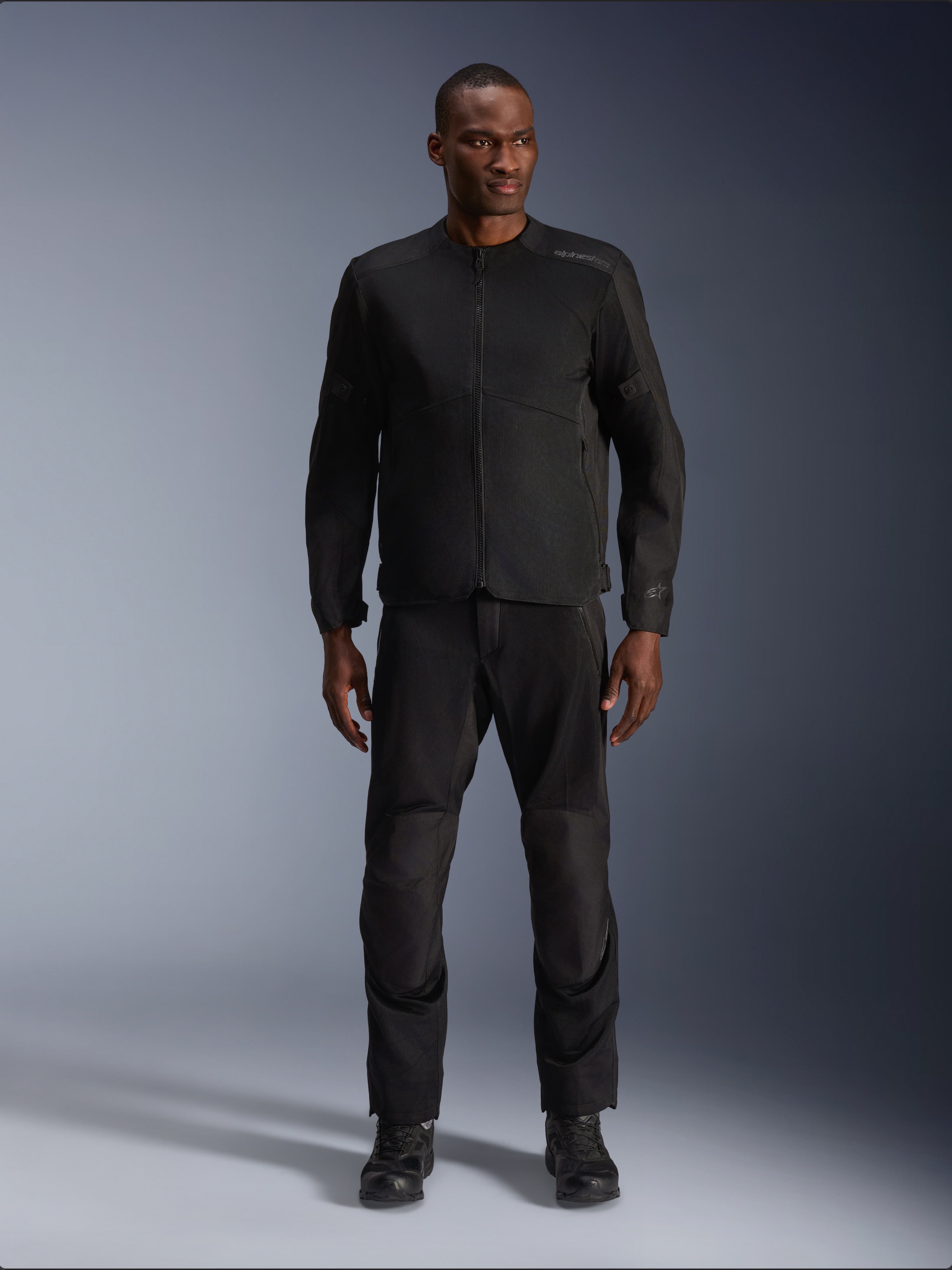 Modelo masculino a usar o Casaco Alpinestars C-1 Air em preto, combinado com calças e botas pretas a condizer, equipamento de motociclismo urbano concebido para deslocações diárias, apresentando uma estética simples com painéis de malha e logótipos subtis.