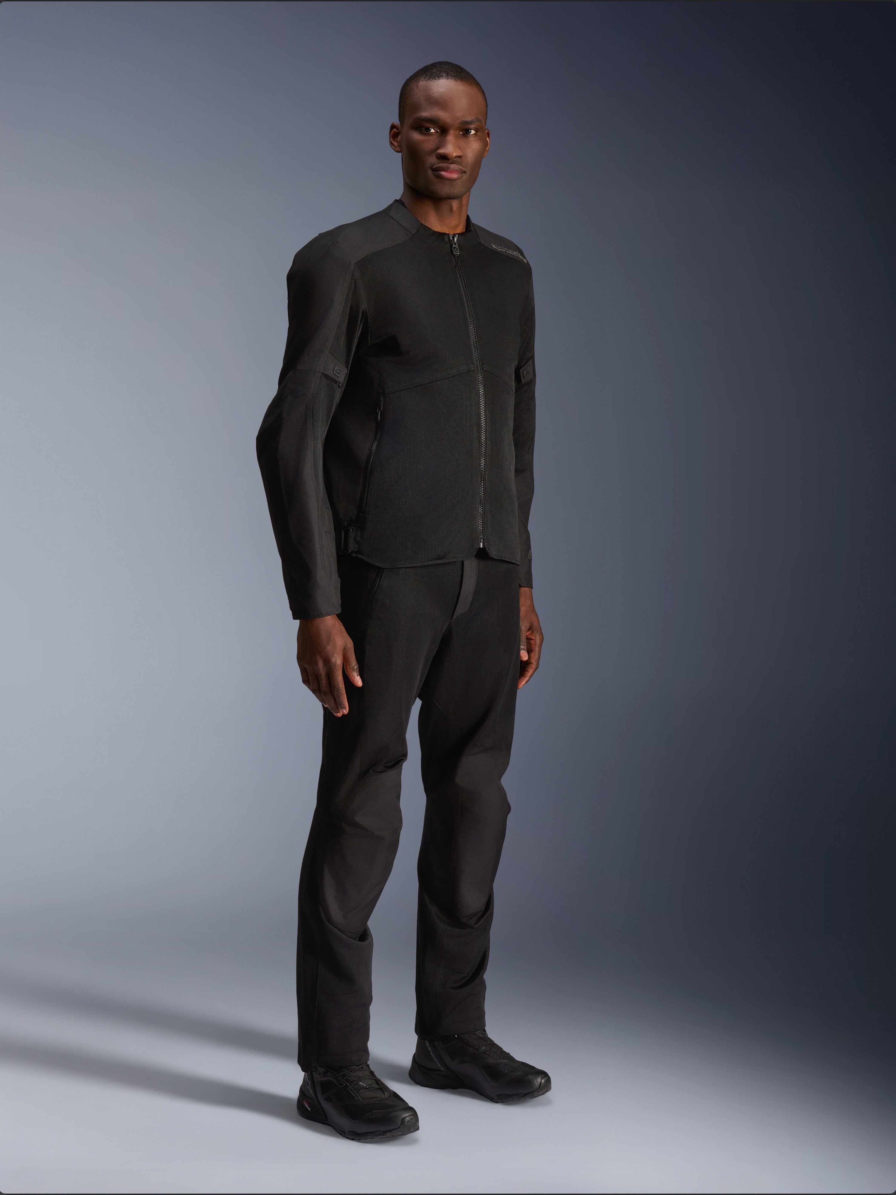 Casaco Alpinestars C-1 Air, preto, vestido por um modelo com calças e sapatos de motociclismo pretos a condizer, com amplos painéis de malha técnica para circulação de ar, estética urbana limpa e um ajuste elegante.