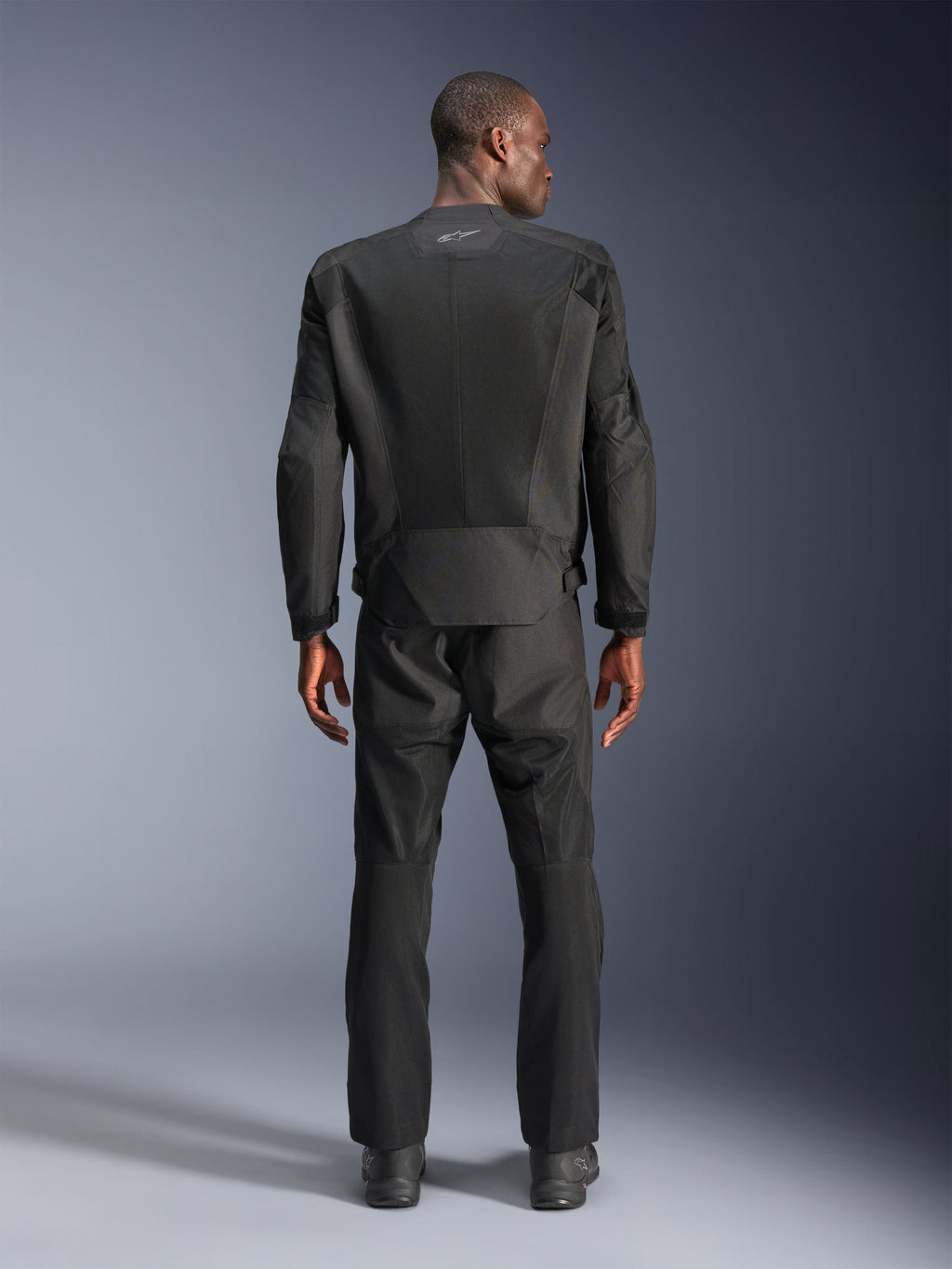 Vista traseira de um homem a usar o casaco Alpinestars C-1 Air em preto, casaco de motociclismo urbano com painéis de malha técnica, calças têxteis e botas pretas a combinar, estética clean com logótipo refletor na parte superior das costas.