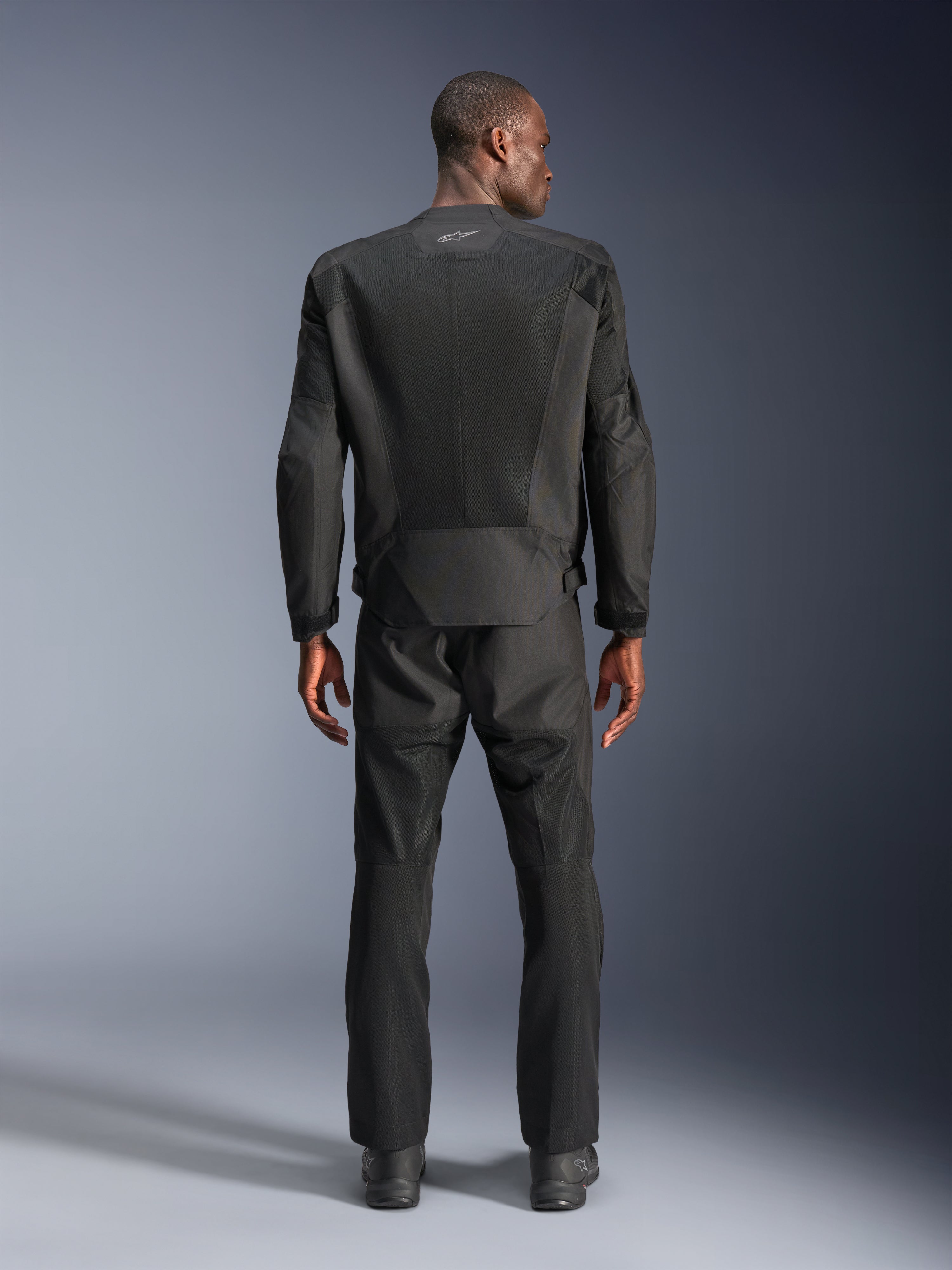 Vista traseira de um homem a usar o casaco Alpinestars C-1 Air em preto, casaco de motociclismo urbano com painéis de malha técnica, calças têxteis e botas pretas a combinar, estética clean com logótipo refletor na parte superior das costas.