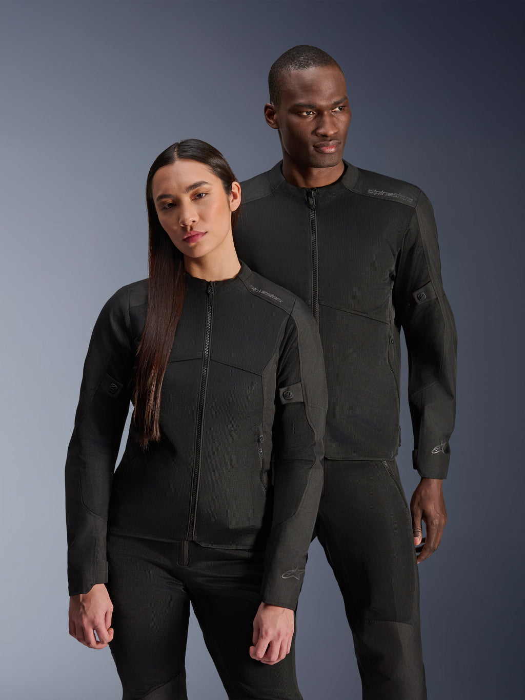 Um homem e uma mulher a usar os casacos Alpinestars C-1 Air e Stella C-1 Air, casacos de motociclismo urbanos em têxtil preto com painéis de malha para ventilação, combinados com calças de condução pretas para um visual limpo e contemporâneo.