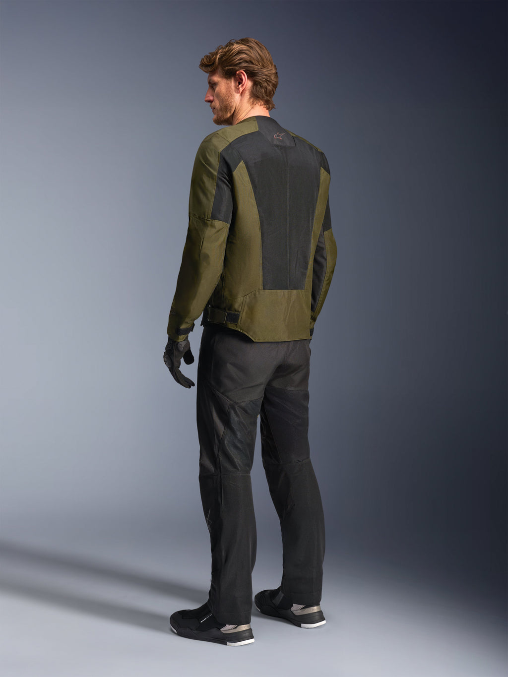 Modelo masculino a usar o Casaco Alpinestars C-1 Air em Khaki, casaco de motociclismo urbano, vista traseira destacando os grandes painéis de malha preta nas costas, combinado com calças técnicas de condução e luvas pretas, equipamento de motociclismo contemporâneo para tempo quente