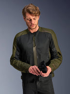 Homem a usar o Casaco Alpinestars C-1 Air em Khaki, casaco de motociclismo urbano com tecido verde azeitona e painéis de malha pretos, a olhar para baixo enquanto segura luvas de motociclismo pretas, estilo limpo e contemporâneo para condução em estrada.