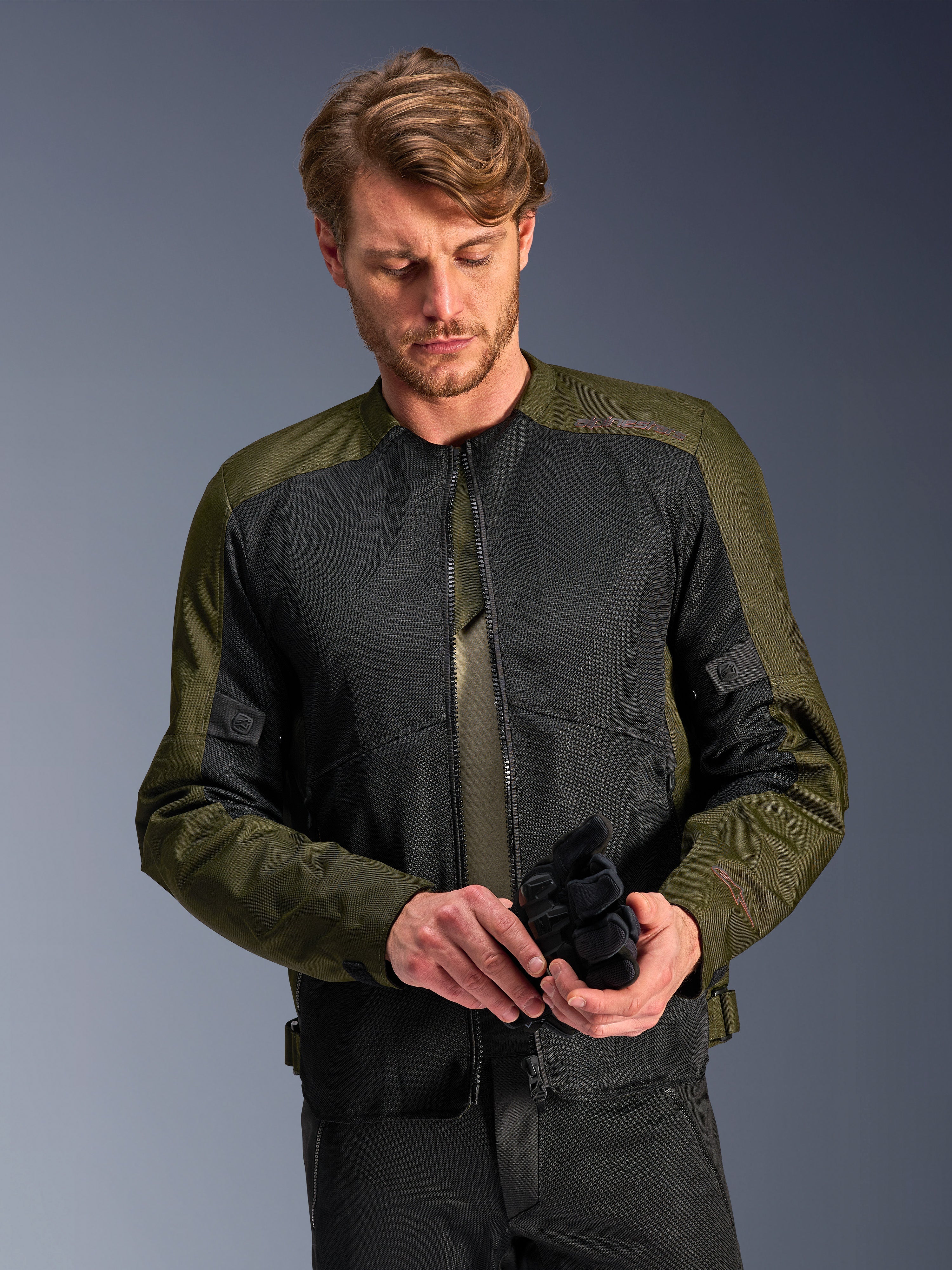 Homem a usar o Casaco Alpinestars C-1 Air em Khaki, casaco de motociclismo urbano com tecido verde azeitona e painéis de malha pretos, a olhar para baixo enquanto segura luvas de motociclismo pretas, estilo limpo e contemporâneo para condução em estrada.