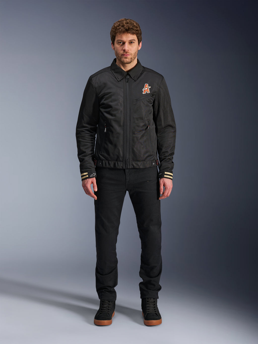 Male model wearing the Alpinestars Ignite Air Jacket in black, featuring a classic shirt collar, full front zipper, and an orange logo on the chest. Styled with black pants and black high-top sneakers for an urban contemporary motorcycle look.