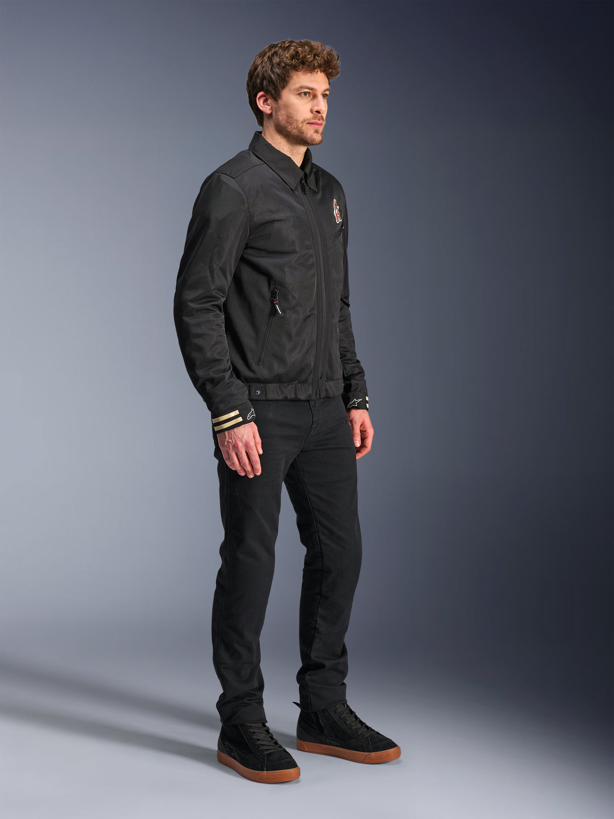 Casaco Alpinestars Ignite Air, casaco de motociclismo urbano, preto, usado por um modelo com calças pretas e sapatilhas pretas, com gola de camisa tradicional, gráficos inspirados em clubes no peito e nos punhos, design contemporâneo e elegante com acabamento em malha e cetim