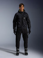 Male model wearing Alpinestars Mogress Air Jacket, black hooded textile jacket with lime punch accents, paired with black utility pants, black gloves, and black and white riding boots, urban motorsport-inspired streetwear design