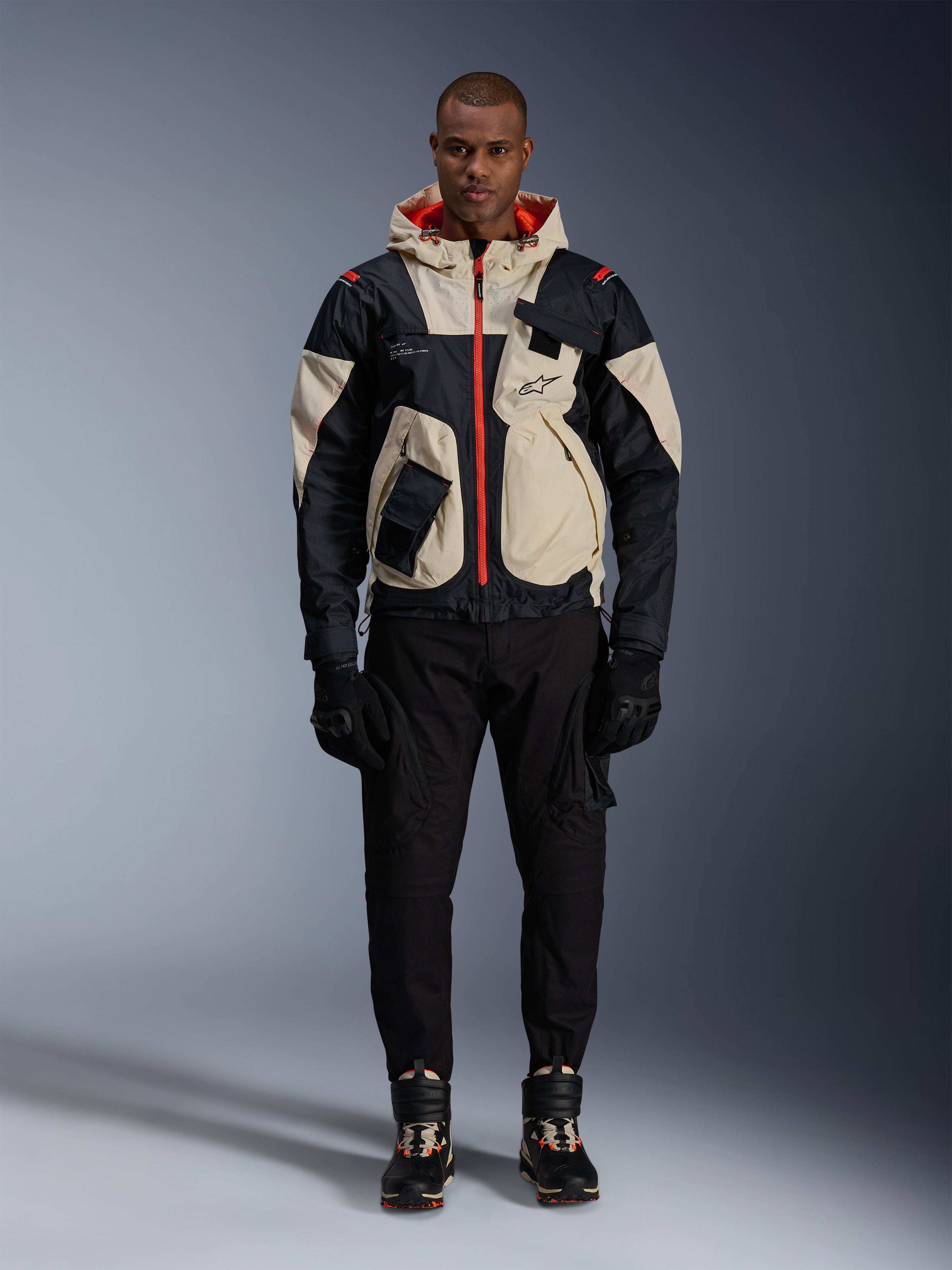 Alpinestars Mogress Air Jacket, Urban Motorcycle Jacket, Black Almond and Hot Orange, worn by a male model with black pants and gloves, utility-inspired design featuring cream panels and bright orange zippers, modern motorsport-influenced streetwear.