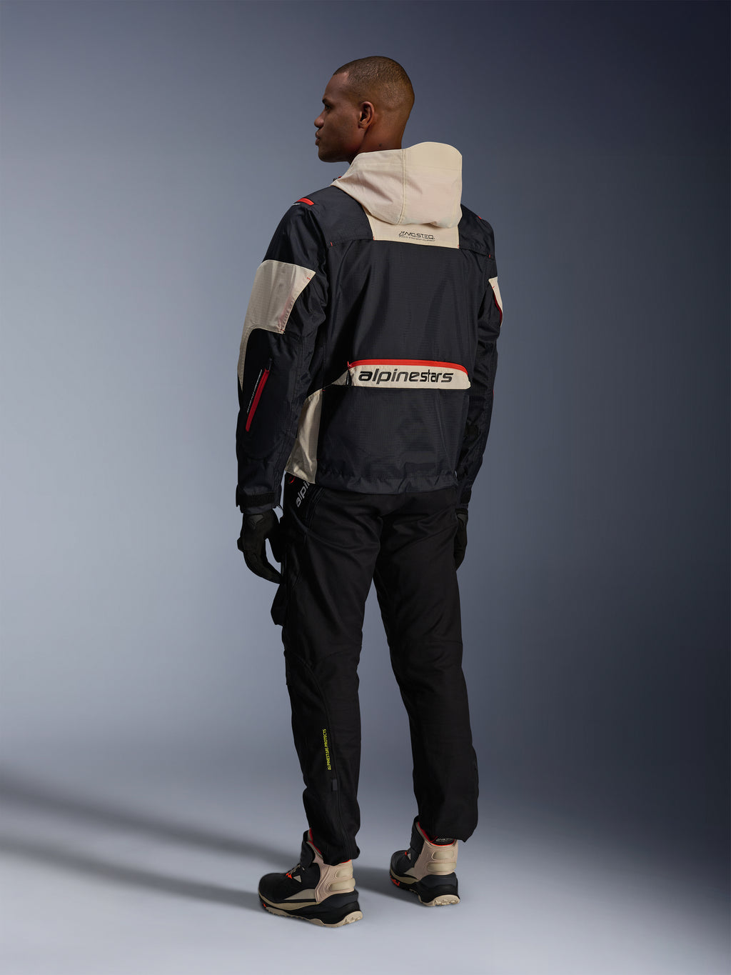 Alpinestars Mogress Air Jacket, Black Almond Hot Orange, back view of rider wearing urban motorcycle gear, almond-colored hood and lower back panel with large Alpinestars branding, orange zip details, paired with black pants and high-top riding boots.