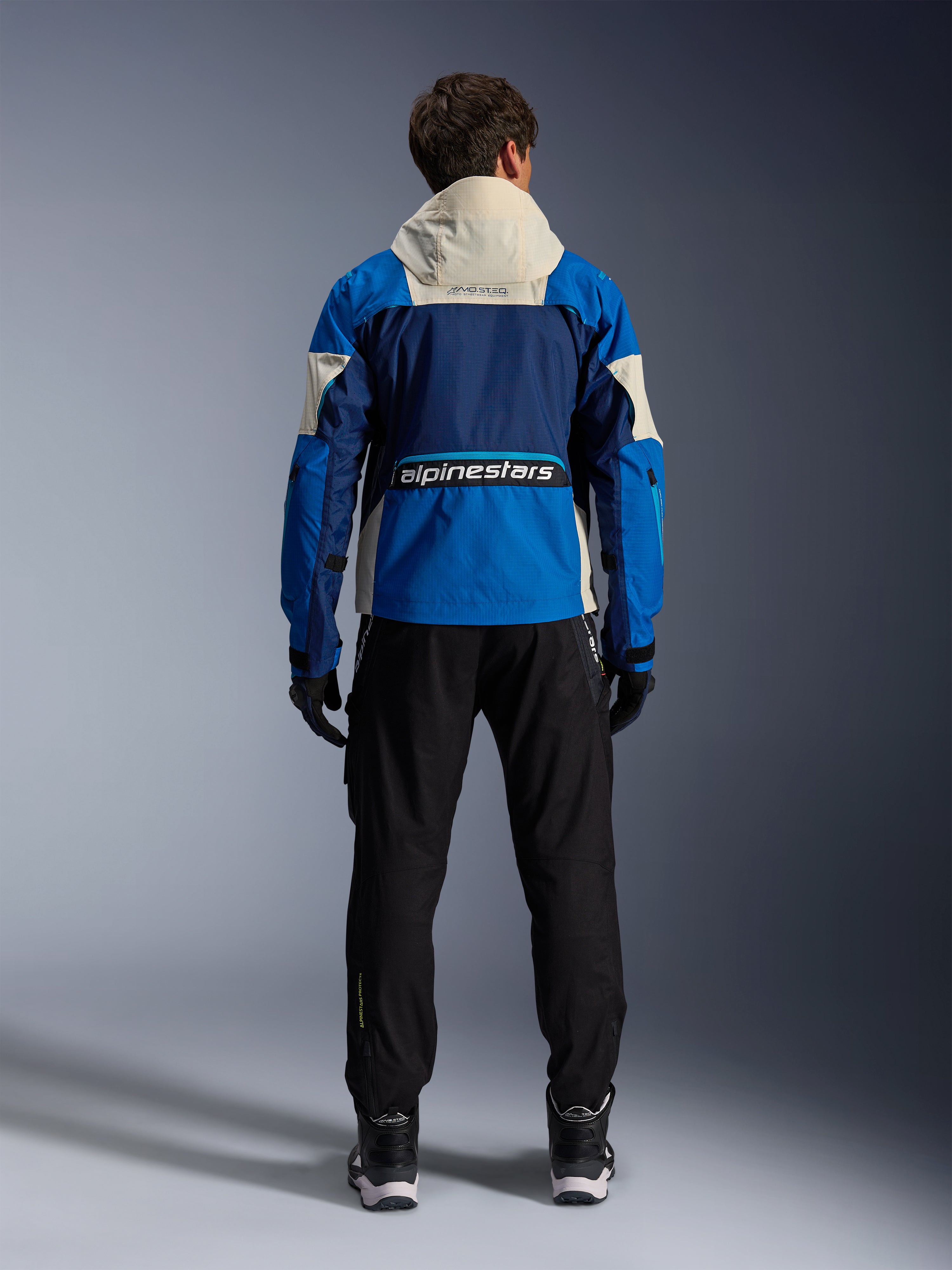 Casaco Alpinestars Mogress Air em Blue Almond, vista traseira de um motociclista a usar o casaco de motociclismo, com painéis azul-marinho e azul brilhante e capuz off-white, logótipo Alpinestars em destaque na parte inferior das costas, estilo de equipamento urbano contemporâneo