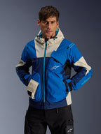 Modelo masculino a usar o Casaco Mogress Air da Alpinestars em Blue Almond, casaco de motociclismo urbano com blocos de cor em azul vivo e azul-marinho, detalhes em creme no capuz e nos bolsos, tecido ripstop técnico com bolsos utilitários, streetwear moderno inspirado no desporto motorizado