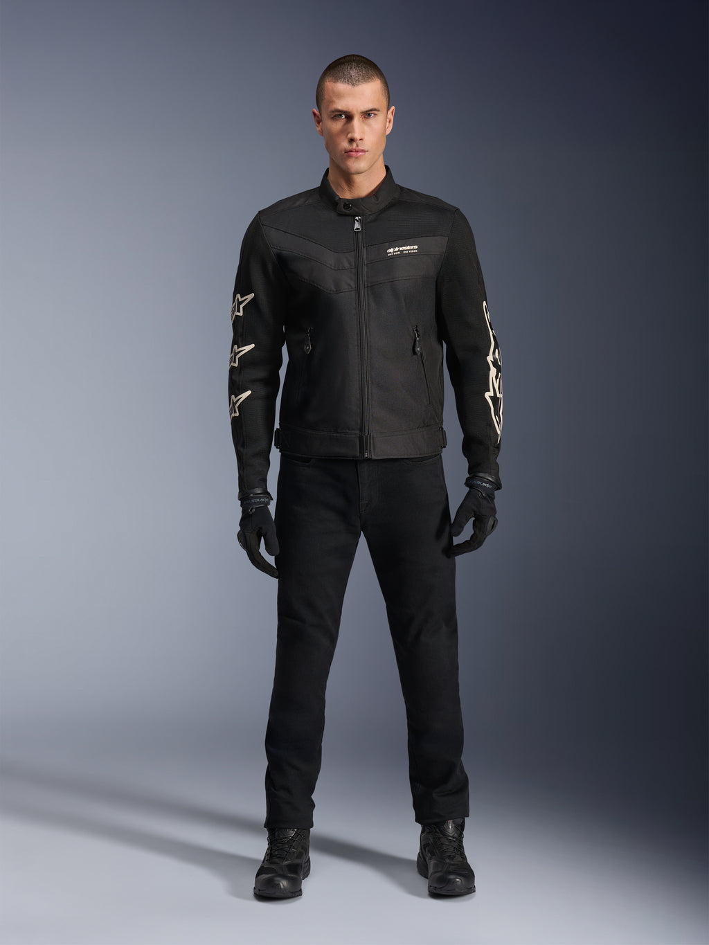 Male model wearing Alpinestars T-Dyno Air Jacket, Black, lightweight textile motorcycle jacket with breathable 3D mesh, white star logos on sleeves, paired with black pants, gloves, and boots, front view standing