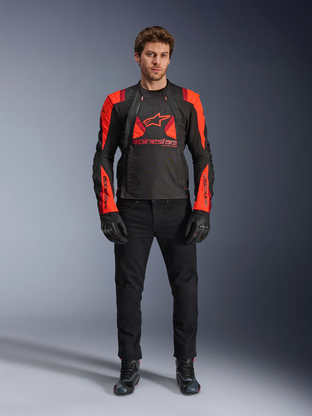 Casaco Alpinestars T-Stunt Air, preto, vermelho fluo e vermelho escuro, usado por um modelo com calças de ganga e luvas de motociclismo pretas, com um design modular 2 em 1 e painéis de malha para fluxo de ar, equipamento de motociclismo com corte desportivo para estrada
