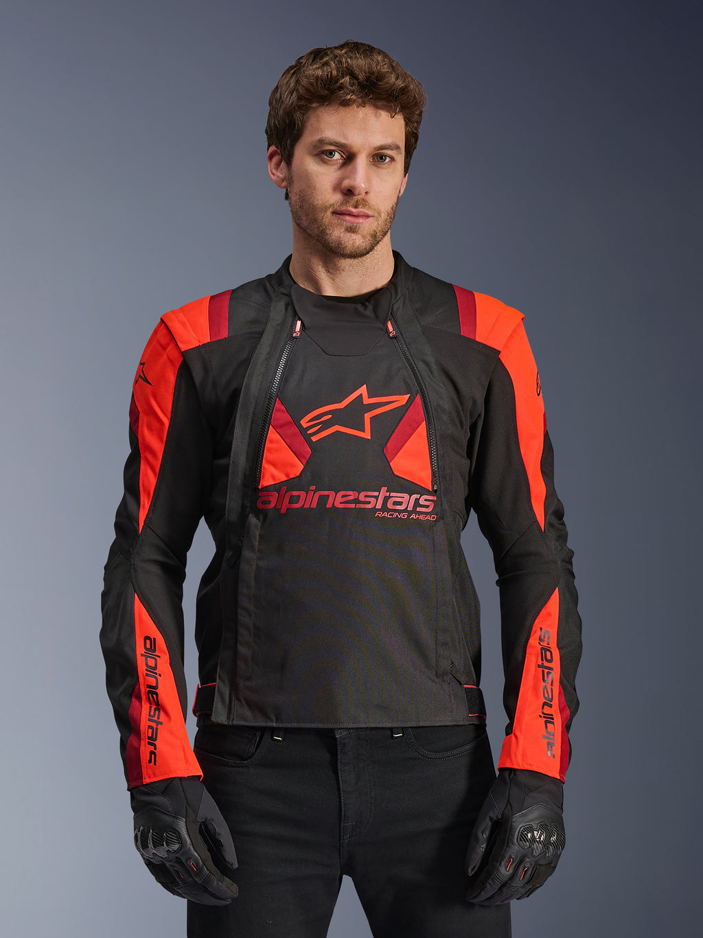 Alpinestars T-Stunt Air Jacket, casaco de motociclismo em tecido, preto, vermelho fluorescente e vermelho escuro, com logótipo de estrela grande e painéis em malha para circulação de ar, usado por modelo com luvas pretas