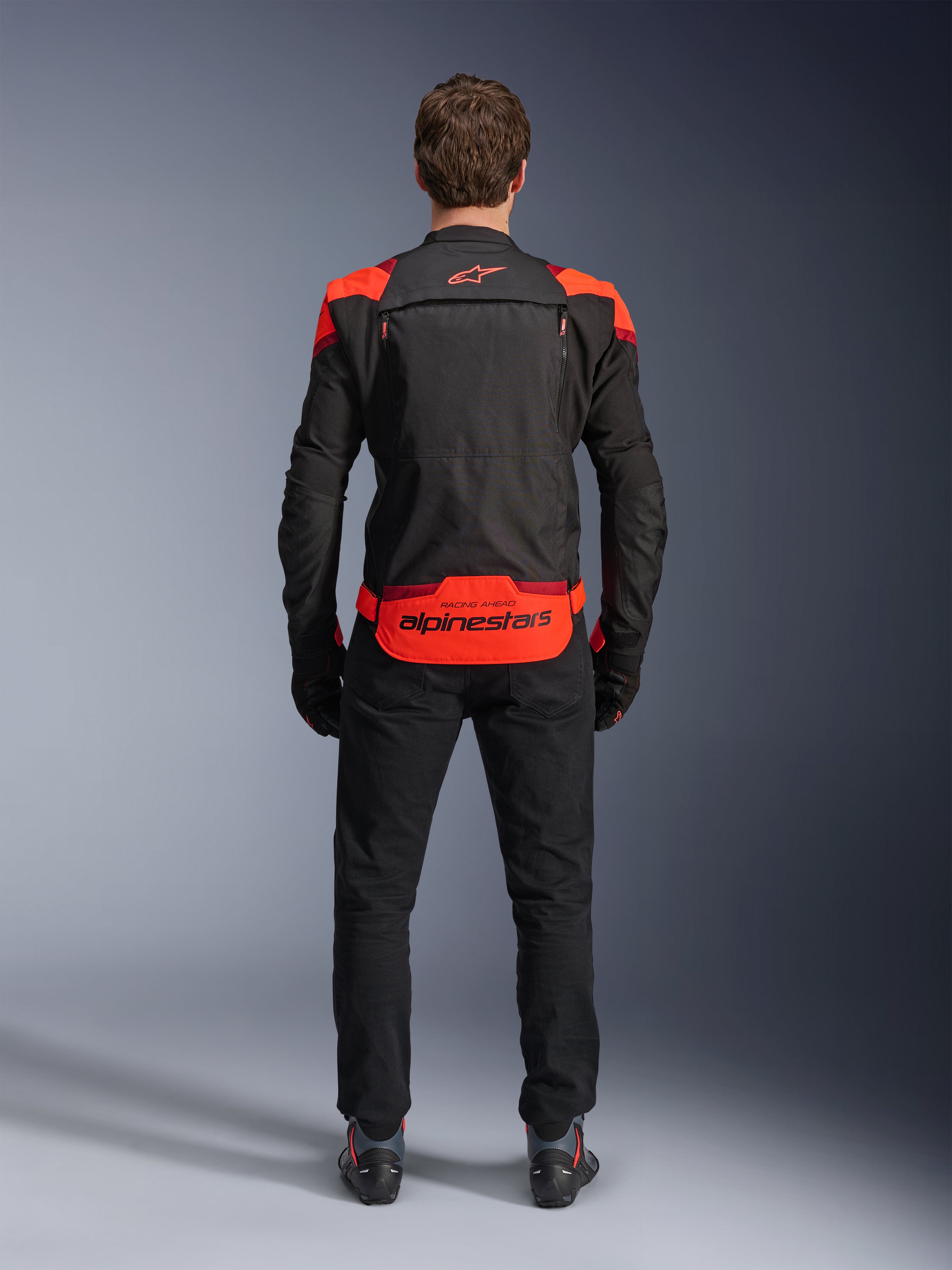Casaco Alpinestars T-Stunt Air, preto com detalhes em vermelho fluo e vermelho escuro, vista traseira no modelo, combinado com calças e botas de condução pretas, com logótipo Alpinestars proeminente no painel inferior traseiro, design modular com construção em malha para ventilação.