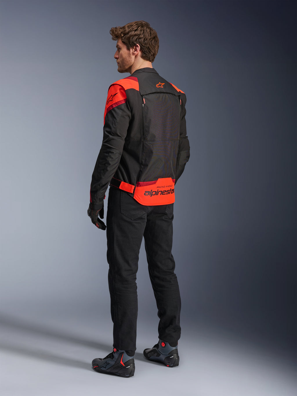 Homem a usar o Casaco Alpinestars T-Stunt Air visto de costas, em Preto Vermelho Fluo e Vermelho Escuro, casaco de motociclismo têxtil com painéis de malha e design de corte desportivo, com o logótipo Alpinestars em destaque na parte inferior das costas, combinado com calças pretas e botas de condução.