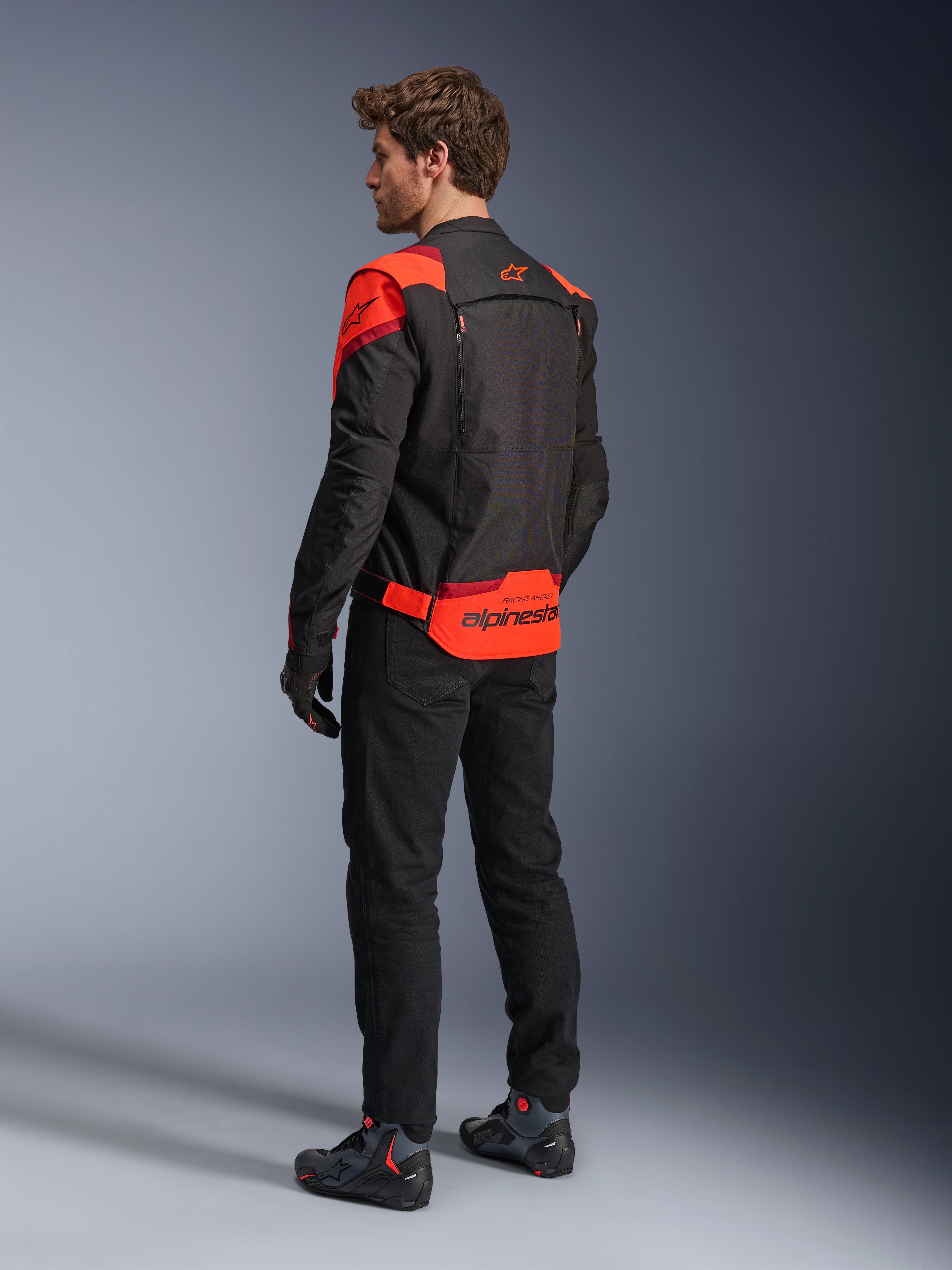 Homem a usar o Casaco Alpinestars T-Stunt Air visto de costas, em Preto Vermelho Fluo e Vermelho Escuro, casaco de motociclismo têxtil com painéis de malha e design de corte desportivo, com o logótipo Alpinestars em destaque na parte inferior das costas, combinado com calças pretas e botas de condução.