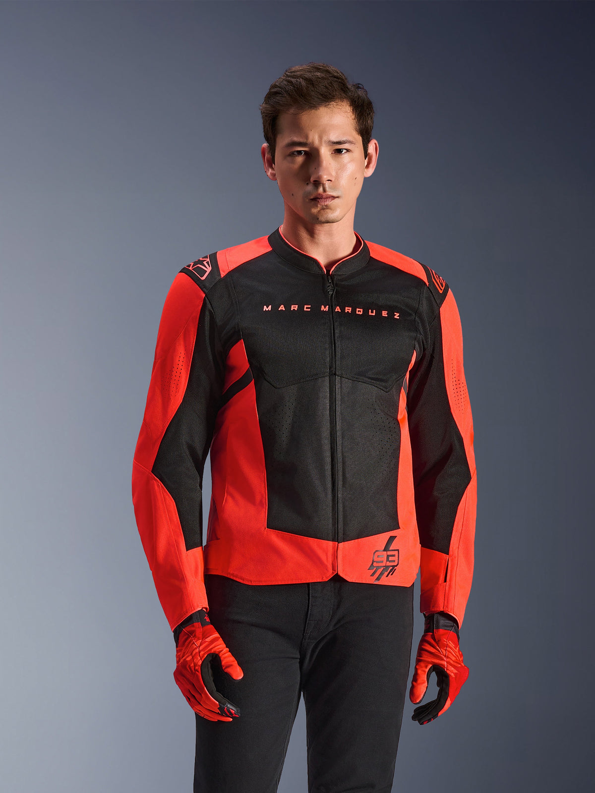 Casaco Alpinestars MM93 T-SPS Air V2, casaco de motociclismo, vermelho fluorescente e preto, usado por um modelo com luvas vermelhas a condizer e calças pretas, com a marca 'Marc Marquez' no peito e logótipo '93', construção em malha desportiva