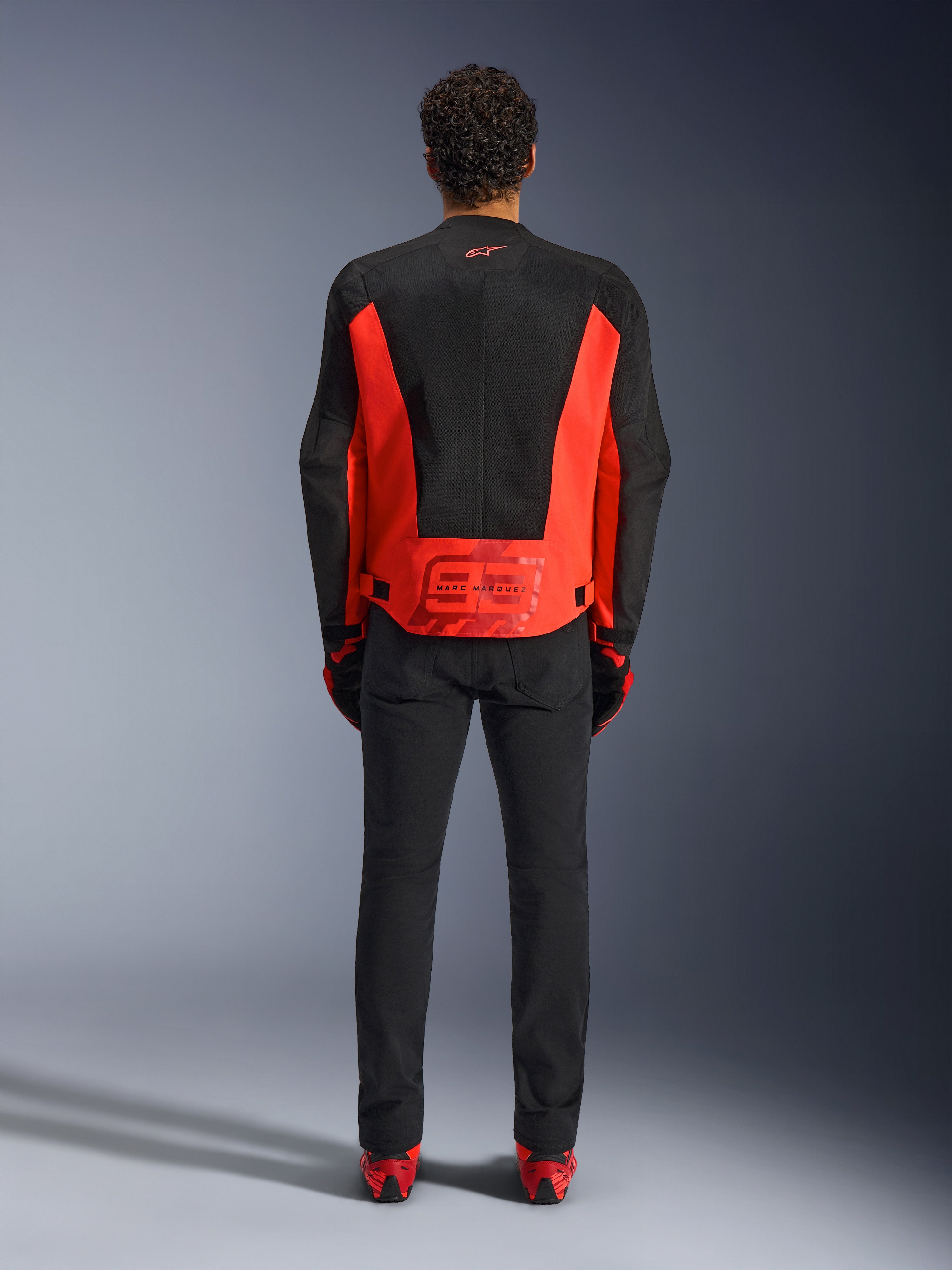 Alpinestars MM93 C-1 Air Jacket, casaco de motociclismo, vermelho fluo e preto, vista traseira com o grafismo Marc Marquez 93 na parte inferior das costas, painéis de malha técnica para ventilação, combinado com calças pretas e equipamento com detalhes em vermelho