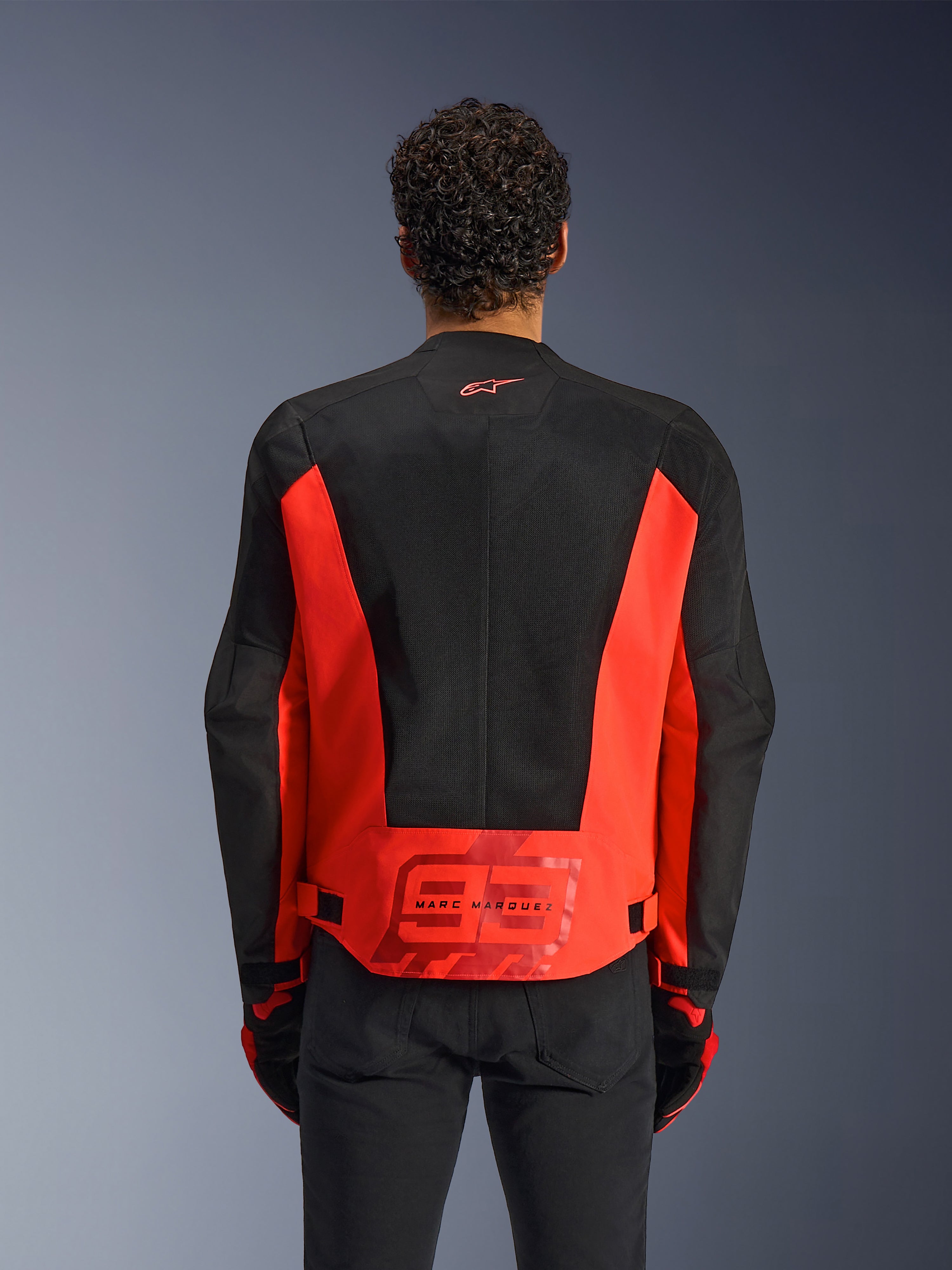 Vista traseira de um homem a usar o casaco Alpinestars MM93 C-1 Air em Vermelho Fluo e Preto, com um logótipo '93' Marc Marquez de grandes dimensões na parte inferior das costas, painéis de malha leve para ventilação, combinado com calças e luvas pretas.