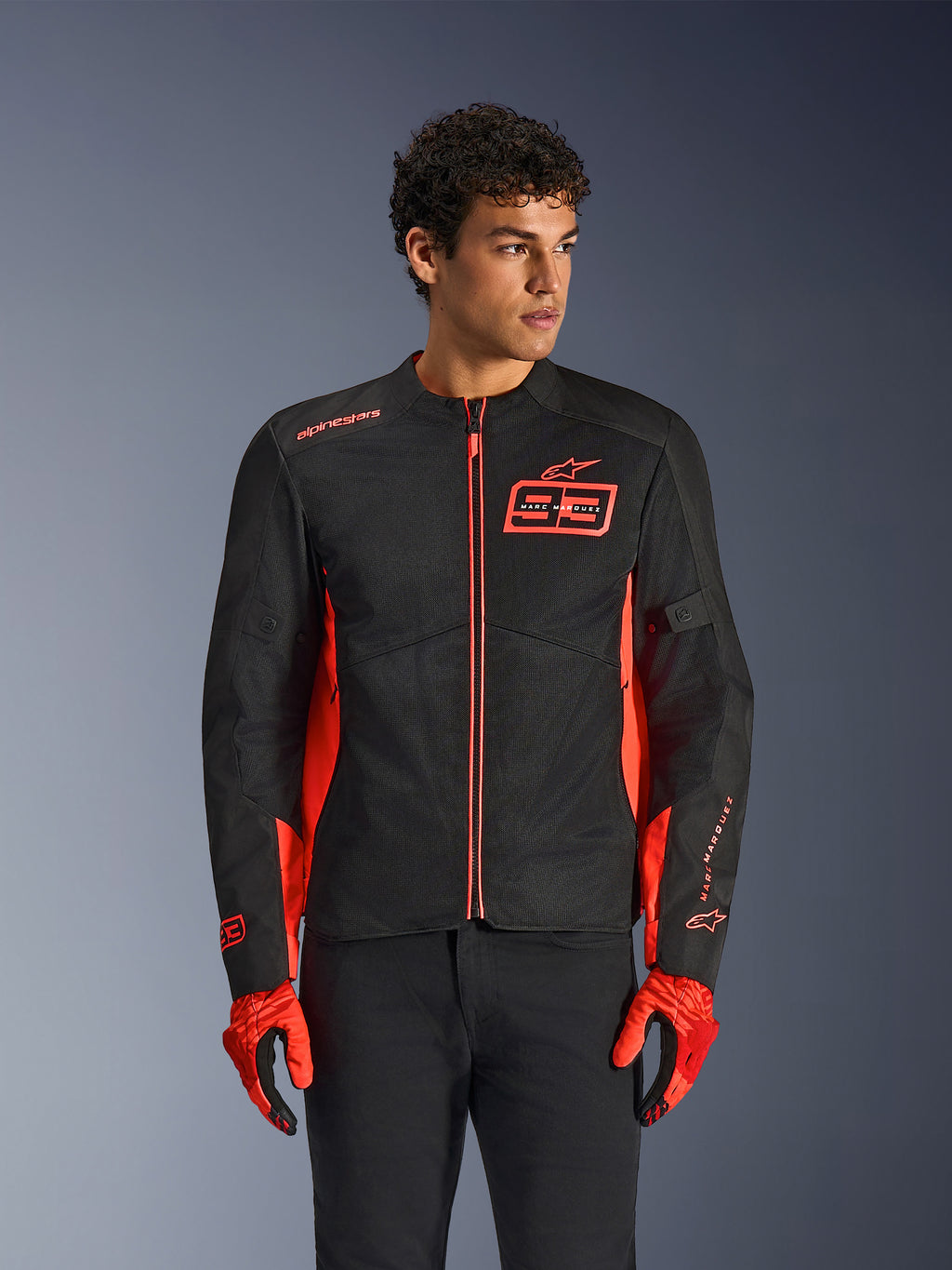 Alpinestars MM93 C-1 Air Jacket, casaco de motociclismo urbano, vermelho fluo e preto, usado por um modelo com luvas vermelhas a condizer e calças pretas, com o logótipo 93 Marc Marquez em destaque no peito e painéis de malha para fluxo de ar