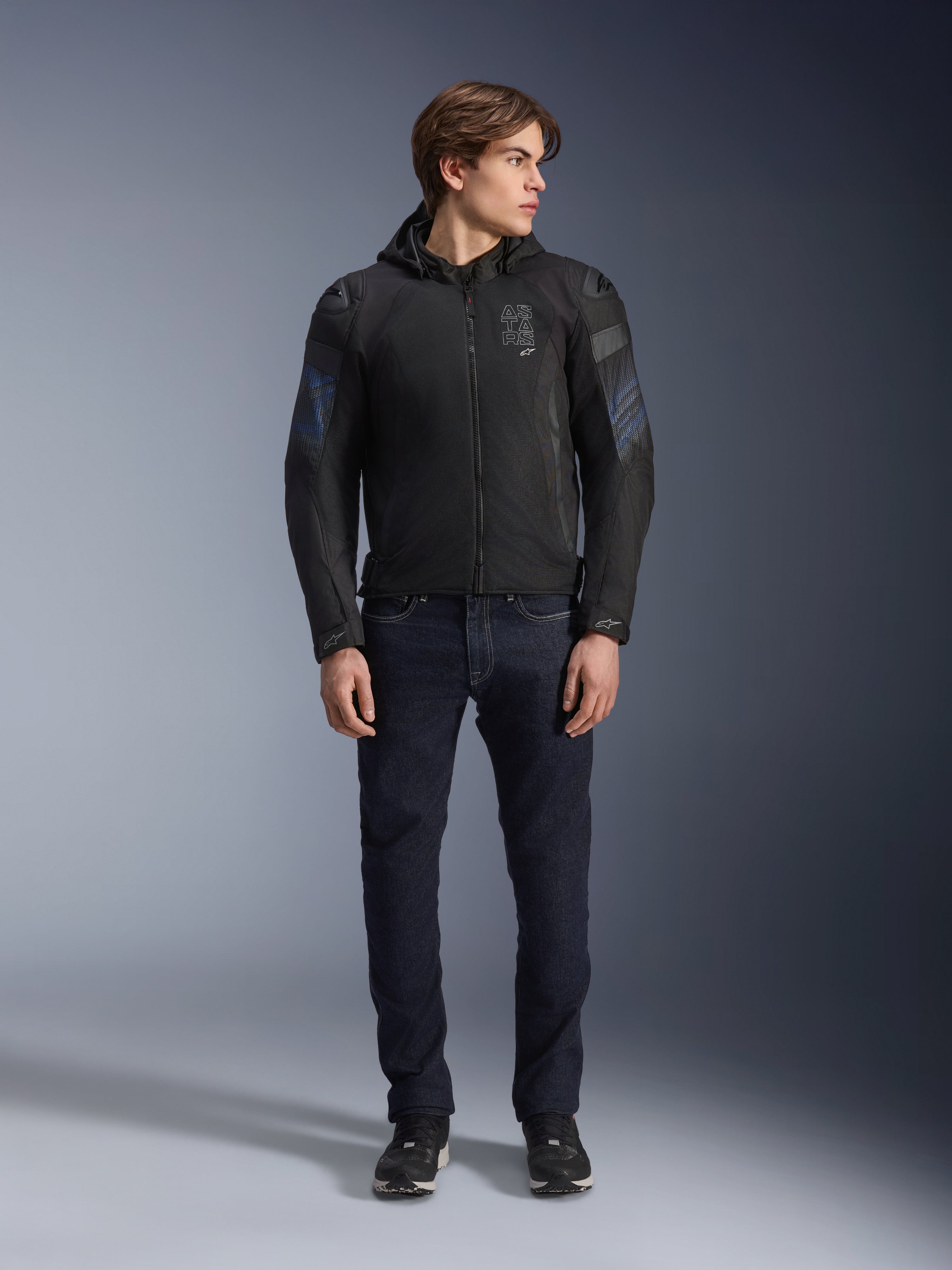 Modelo masculino a usar o casaco Alpinestars Zaca Air Venom WP em preto e azul elétrico, casaco de motociclismo em têxtil com painéis de malha 3D e capuz amovível, combinado com calças de ganga escura, equipamento de condução moderno de inspiração desportiva