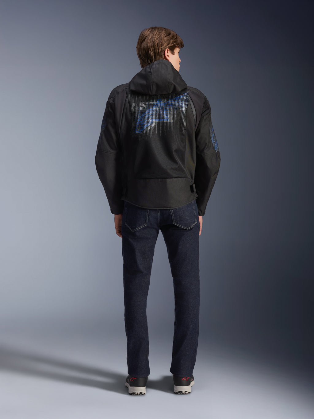 Casaco Alpinestars Zaca Air Venom WP, preto e azul elétrico, vista traseira com um grande logótipo Alpinestars azul no painel de malha das costas, capuz removível, usado com calças de ganga escuras e sapatilhas, equipamento de motociclismo desportivo para todas as condições climatéricas