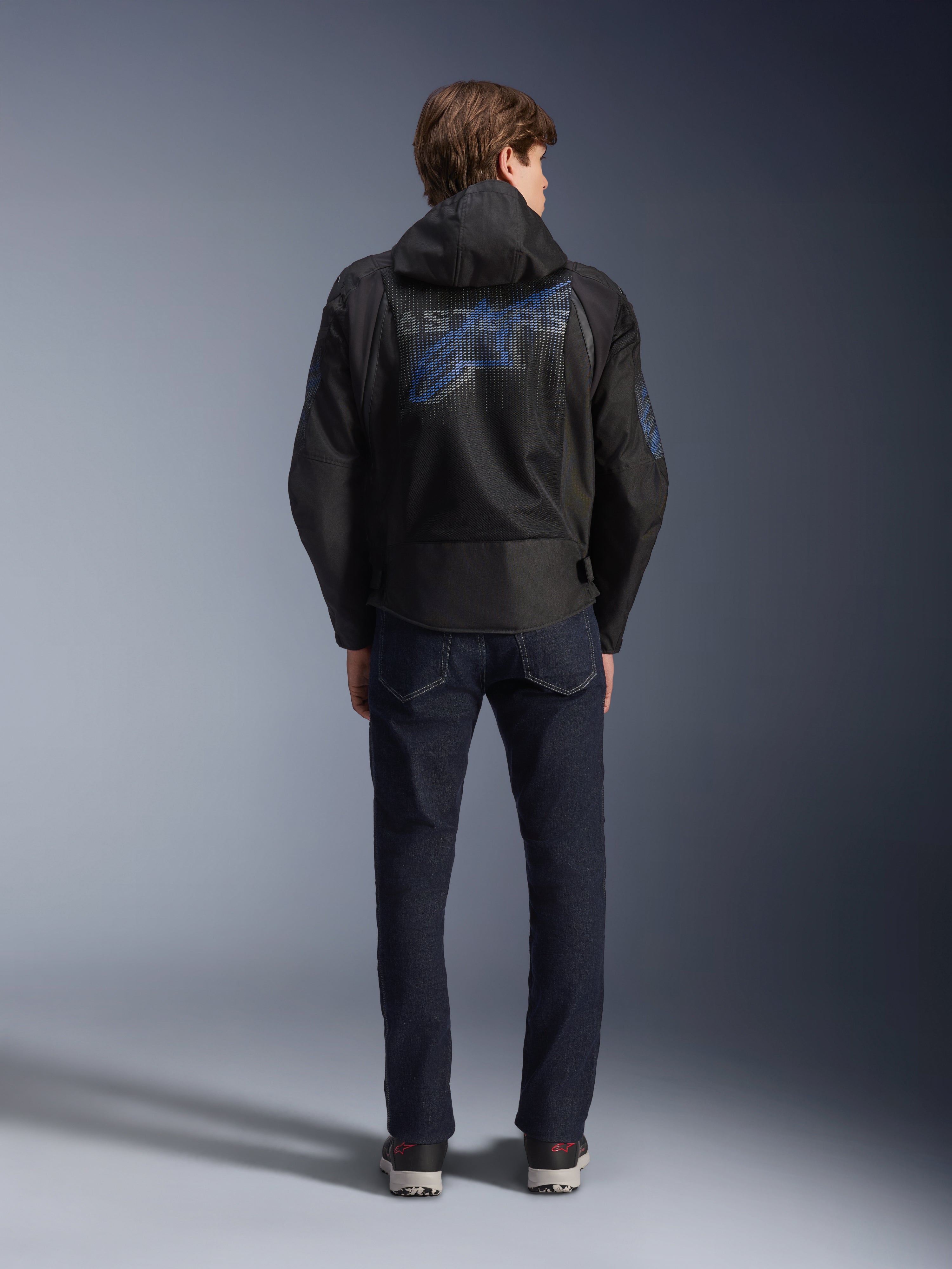 Casaco Alpinestars Zaca Air Venom WP, preto e azul elétrico, vista traseira com um grande logótipo Alpinestars azul no painel de malha das costas, capuz removível, usado com calças de ganga escuras e sapatilhas, equipamento de motociclismo desportivo para todas as condições climatéricas