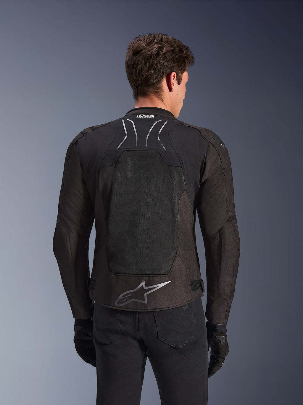 Casaco Alpinestars SMX Air em Black Anthracite, vista traseira num modelo, com um grande painel traseiro em malha técnica para ventilação, marca Tech-Air Ready na gola e logótipo Astar prateado na parte inferior das costas, combinado com luvas pretas e calças de condução