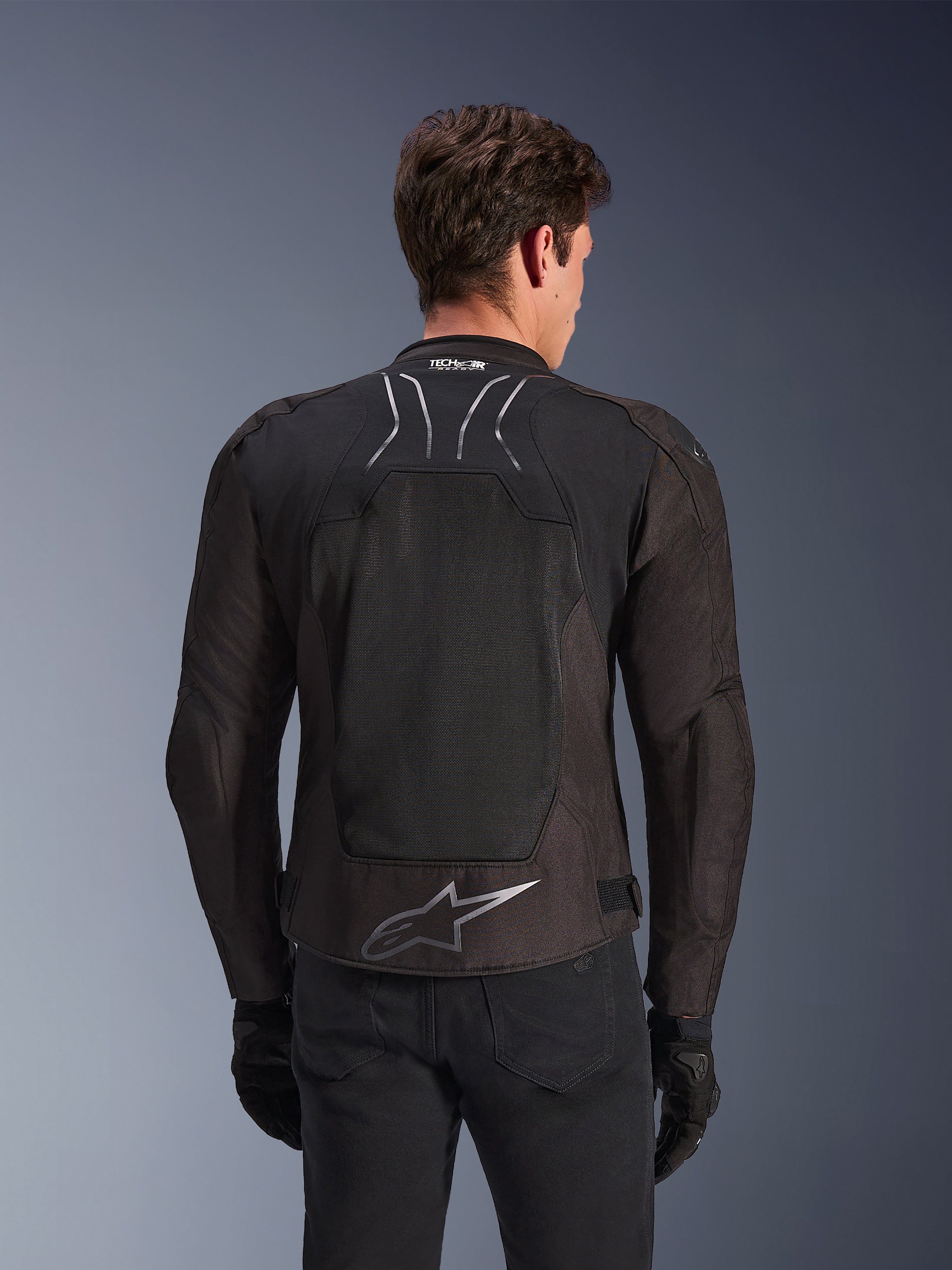 Casaco Alpinestars SMX Air em Black Anthracite, vista traseira num modelo, com um grande painel traseiro em malha técnica para ventilação, marca Tech-Air Ready na gola e logótipo Astar prateado na parte inferior das costas, combinado com luvas pretas e calças de condução