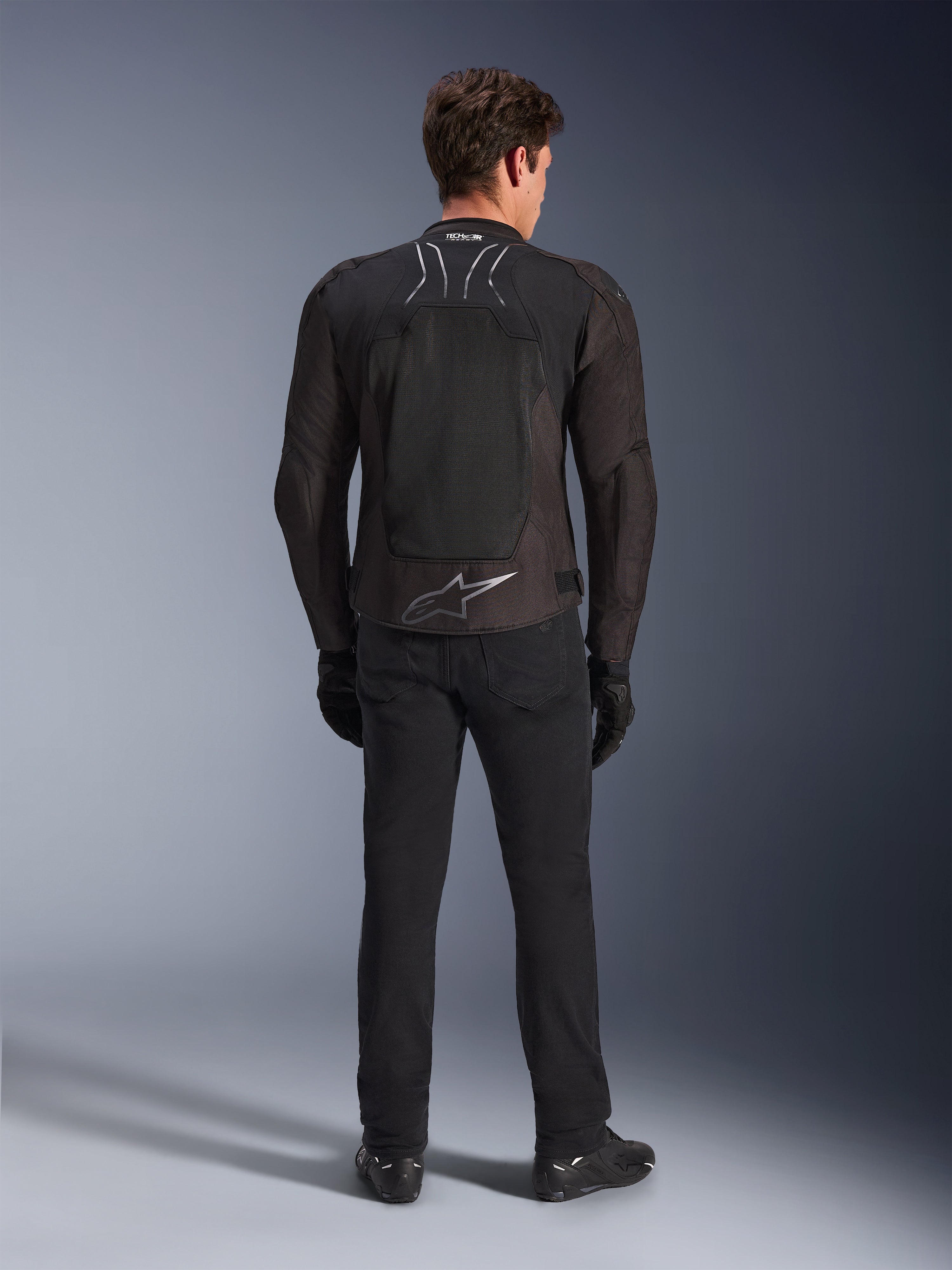 Casaco Alpinestars SMX Air, Preto Antracite, vista traseira num modelo masculino, com um grande logótipo Alpinestars prateado na parte inferior das costas e painéis de malha de alto fluxo, usado com calças de condução, luvas e botas pretas, equipamento de motociclismo desportivo