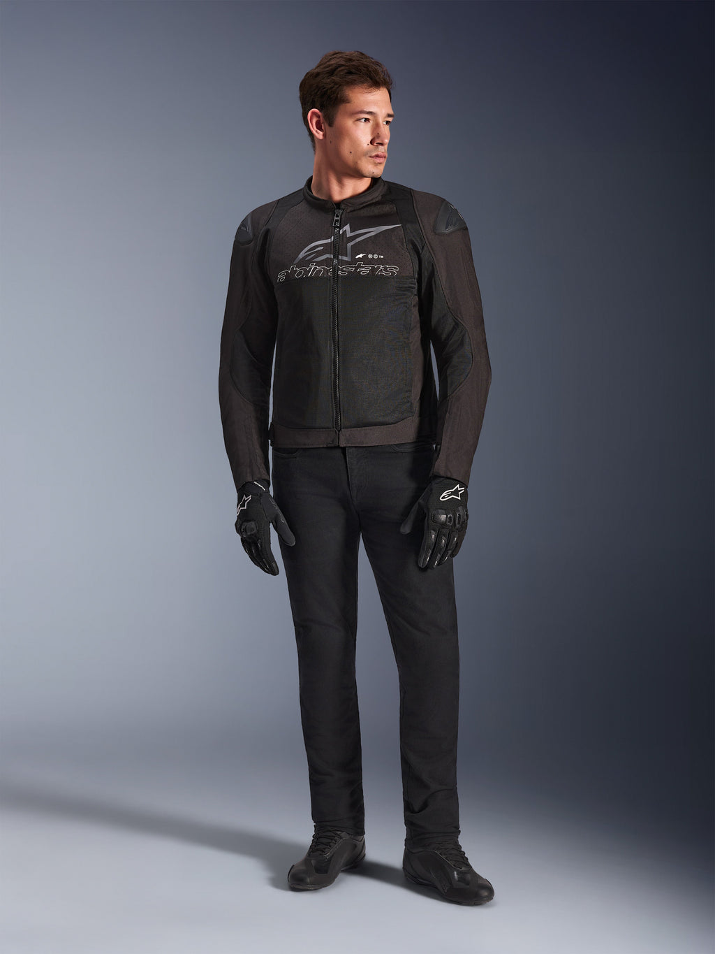 Modelo masculino a vestir o Casaco Alpinestars SMX Air, casaco de motociclismo Black Anthracite com painéis de malha ventilada e logótipo cinzento no peito, combinado com calças pretas e luvas de proteção, num cenário de estúdio com fundo gradiente