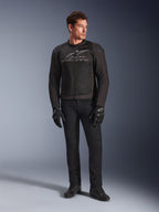Modelo masculino a vestir o Casaco Alpinestars SMX Air, casaco de motociclismo Black Anthracite com painéis de malha ventilada e logótipo cinzento no peito, combinado com calças pretas e luvas de proteção, num cenário de estúdio com fundo gradiente