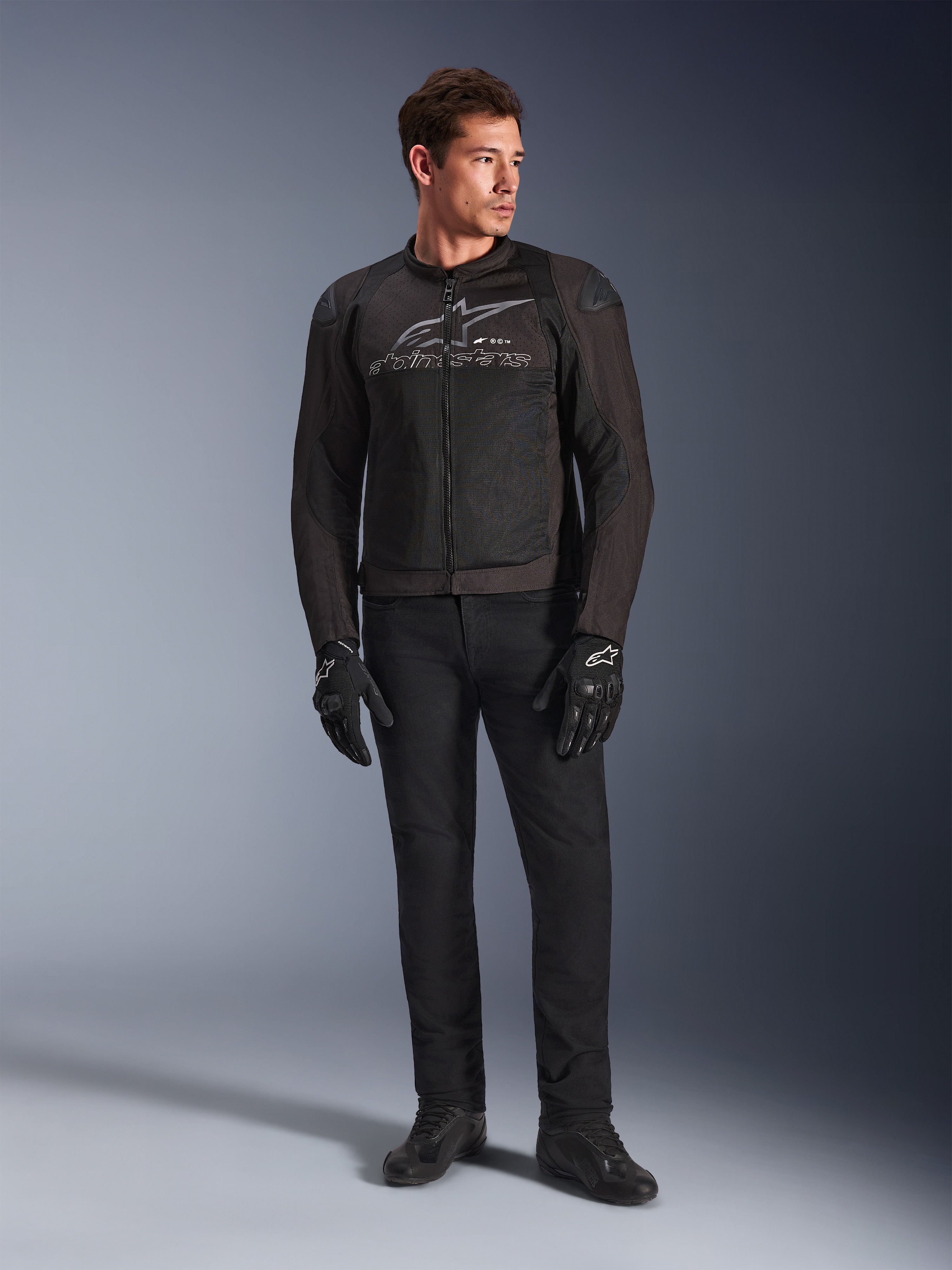 Modelo masculino a vestir o Casaco Alpinestars SMX Air, casaco de motociclismo Black Anthracite com painéis de malha ventilada e logótipo cinzento no peito, combinado com calças pretas e luvas de proteção, num cenário de estúdio com fundo gradiente
