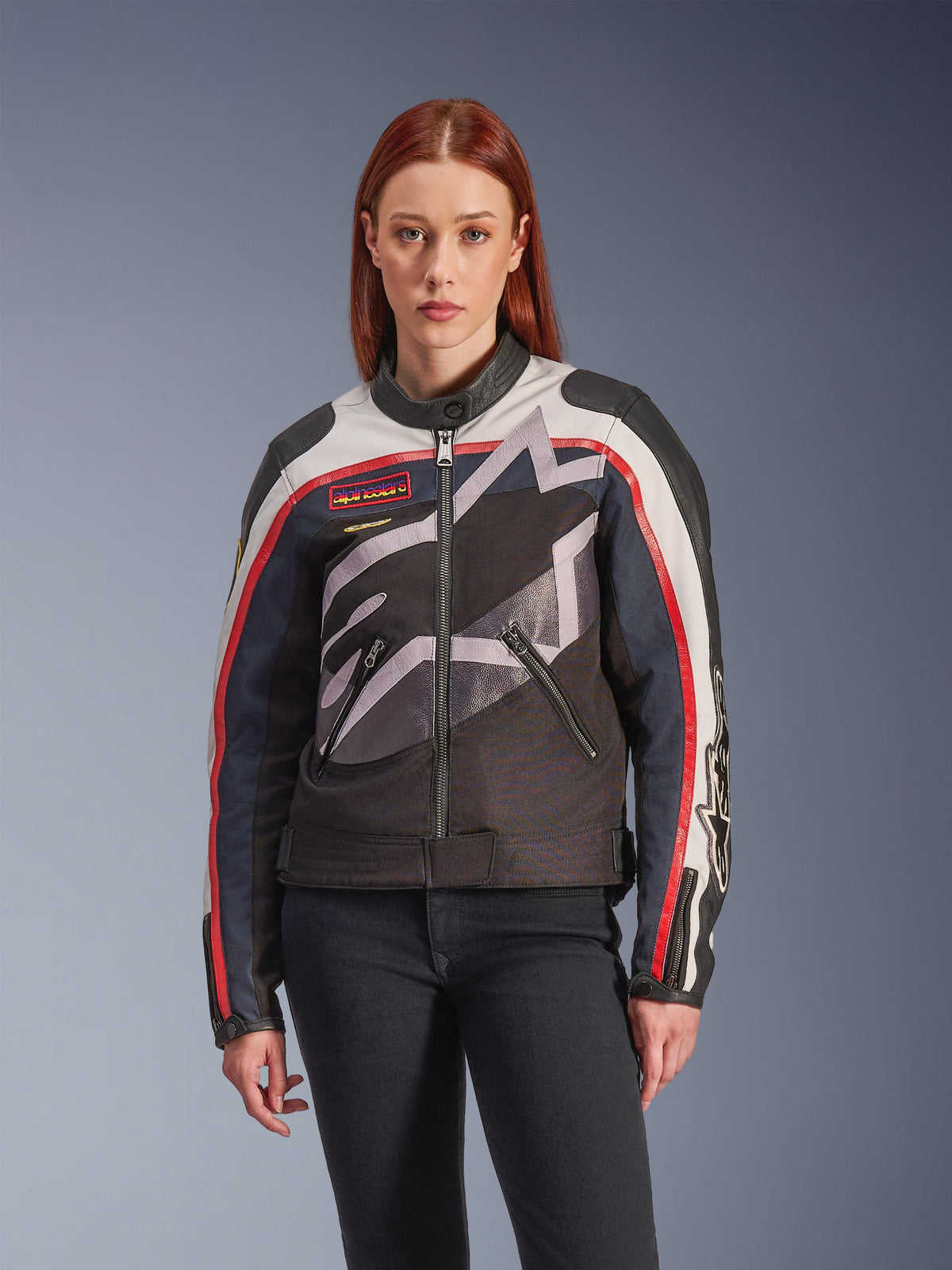 Casaco Alpinestars Idles, casaco de motociclismo feminino, off-white, vermelho flama e preto, com um grande logótipo de estrela estilizado, emblemas no peito de inspiração vintage, gola alta de motociclista, bolsos com fecho de correr e mangas com riscas, usado por uma modelo com calças de ganga escuras.