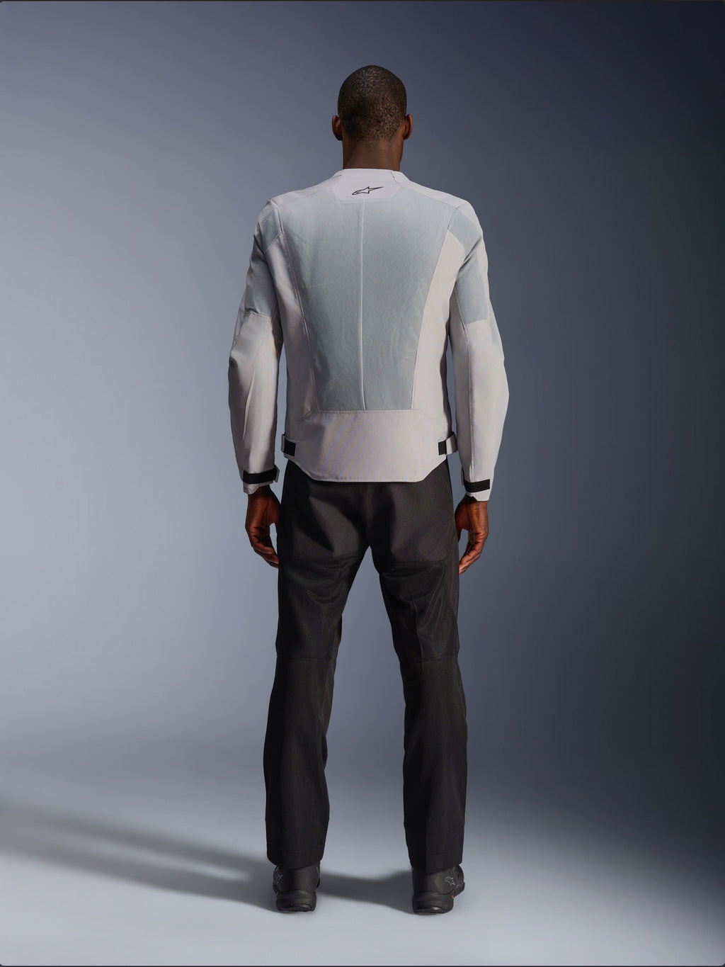 Casaco Alpinestars C-1 Air em Silver, vista traseira num modelo masculino, casaco de motociclismo têxtil com grandes painéis de ventilação em malha técnica, pequeno logótipo preto na gola e ajustadores de cintura, usado com calças de condução pretas para um visual urbano contemporâneo.