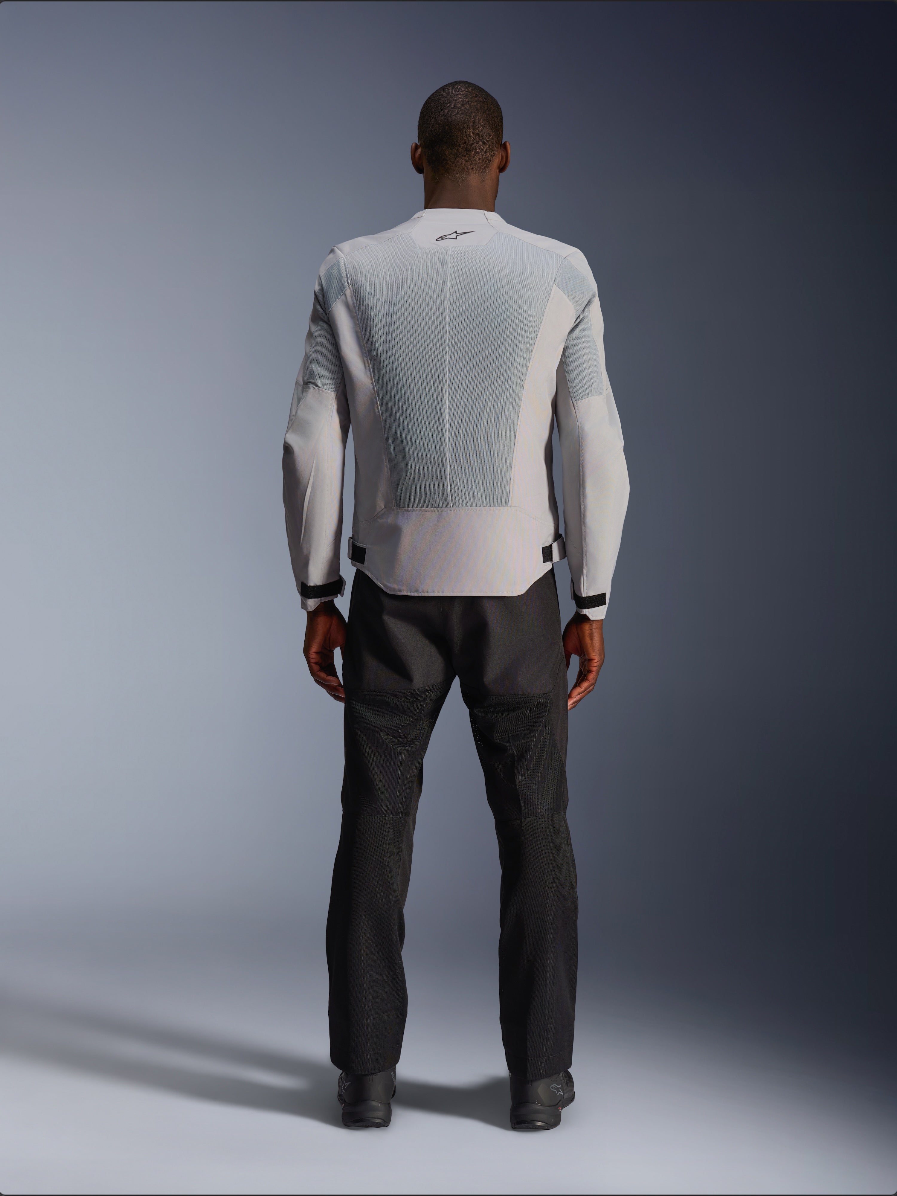 Casaco Alpinestars C-1 Air em Silver, vista traseira num modelo masculino, casaco de motociclismo têxtil com grandes painéis de ventilação em malha técnica, pequeno logótipo preto na gola e ajustadores de cintura, usado com calças de condução pretas para um visual urbano contemporâneo.
