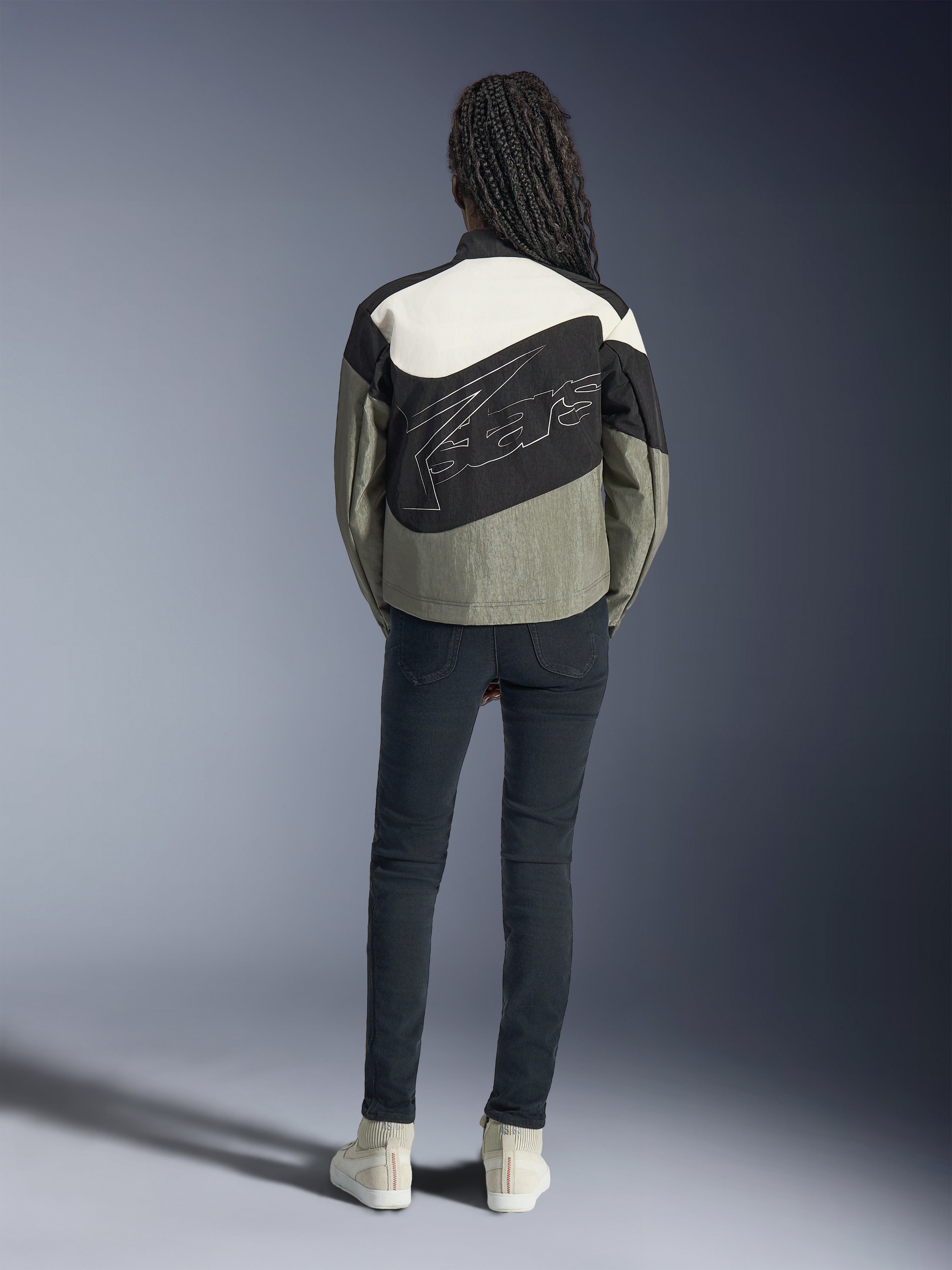 Rear view of a model wearing the Alpinestars Stella Moshell Jacket in Black, Gray, and Off-White, featuring an asymmetrical design and large stylized outline logo on the back, paired with dark slim-fit jeans and white sneakers, urban streetwear-inspired motorcycle apparel for women.