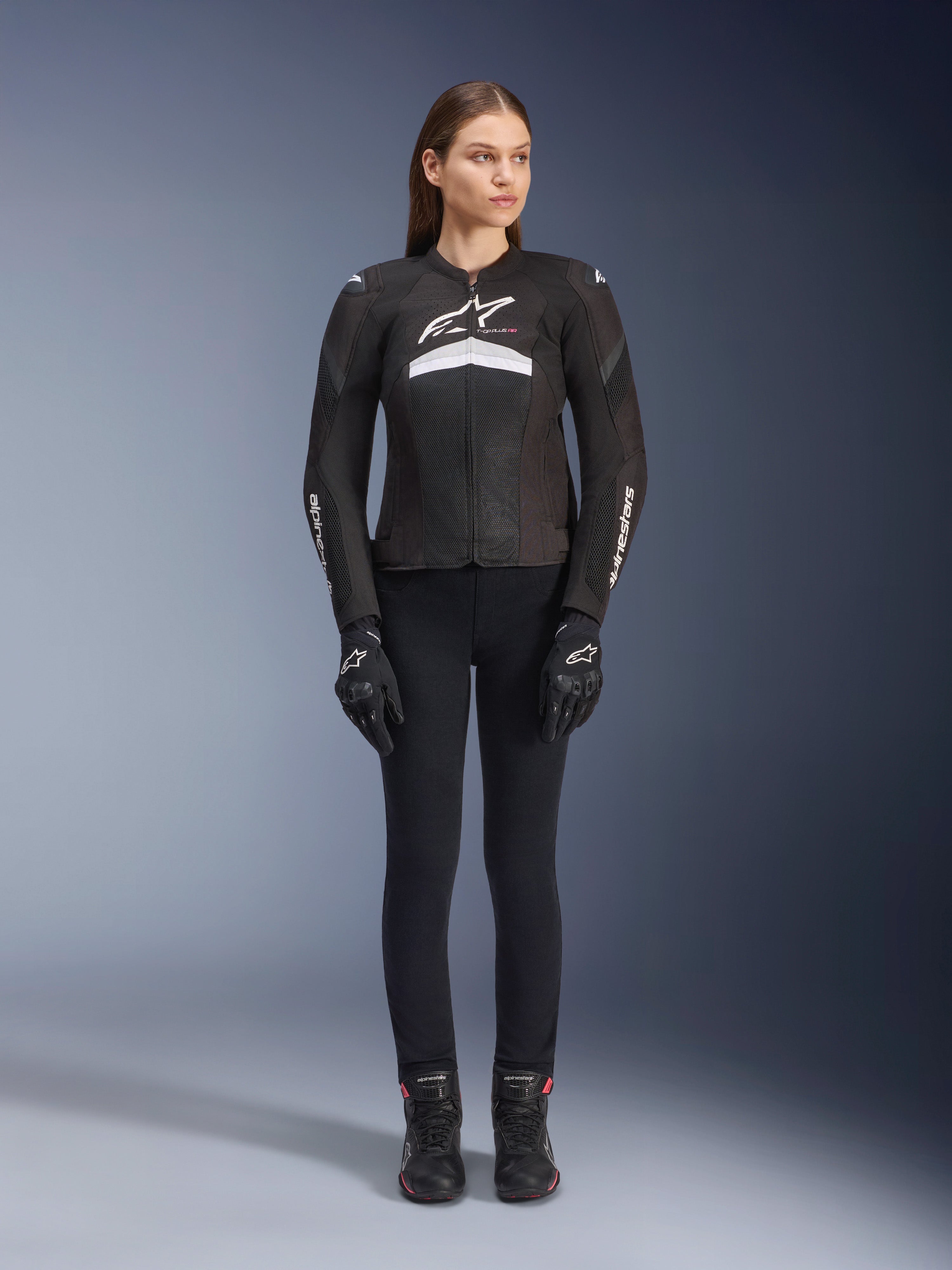 Female rider wearing the Stella T-GP Plus R V4 Airflow Jacket, Black and White, paired with black riding pants, gloves, and boots, featuring mesh panels for ventilation and white Alpinestars branding on the chest and arms