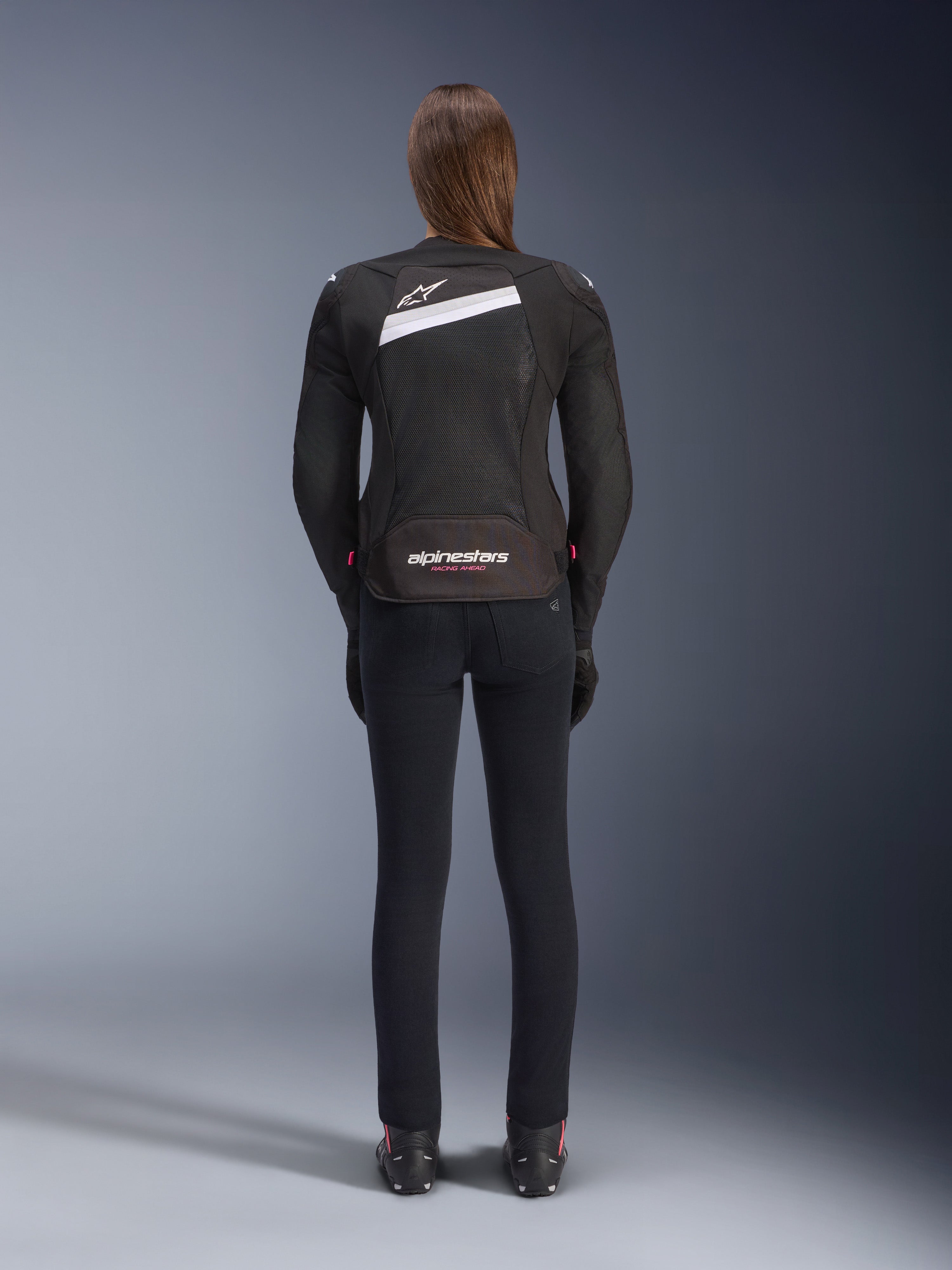 Alpinestars Stella T-GP Plus R V4 Airflow Jacket, Black and White, rear view showing extensive mesh ventilation panels and Alpinestars branding, worn by a female rider with slim-fit black motorcycle pants and boots, sport-fit textile gear for women