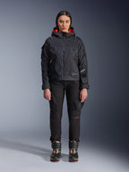 Female model wearing the Alpinestars Molly Air Jacket in Black and Fluo Coral, featuring a utility-inspired design with a hood and coral inner lining, paired with black riding pants and motorcycle boots, urban contemporary motorcycle gear
