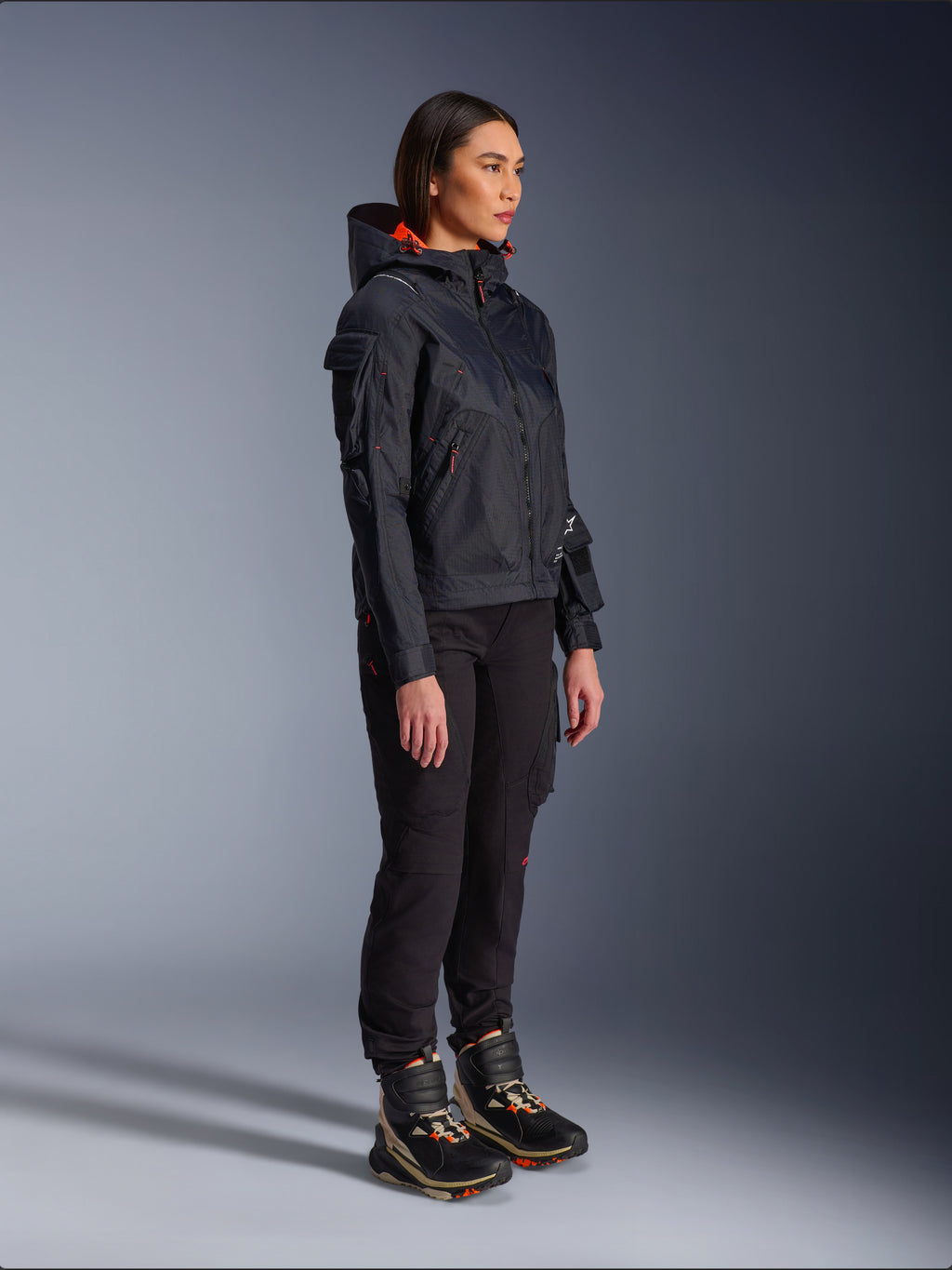 Female rider wearing the Alpinestars Molly Air Jacket in Black Fluo Coral, lightweight textile motorcycle jacket with a bright orange hood lining, styled with black riding pants and technical boots, urban contemporary utility-inspired design.